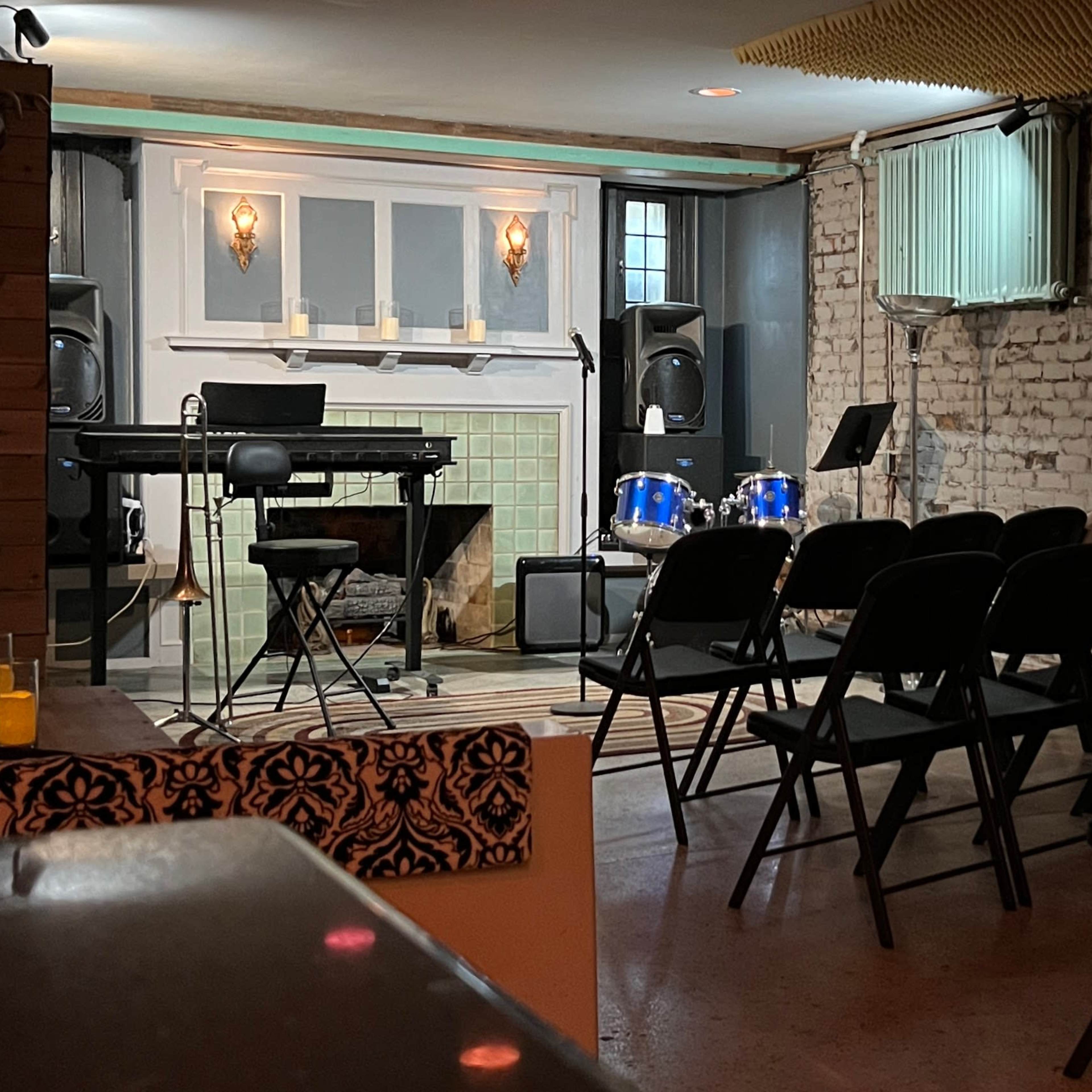 A small, empty performance space featuring a keyboard, drums, and rows of folding chairs arranged for an audience.