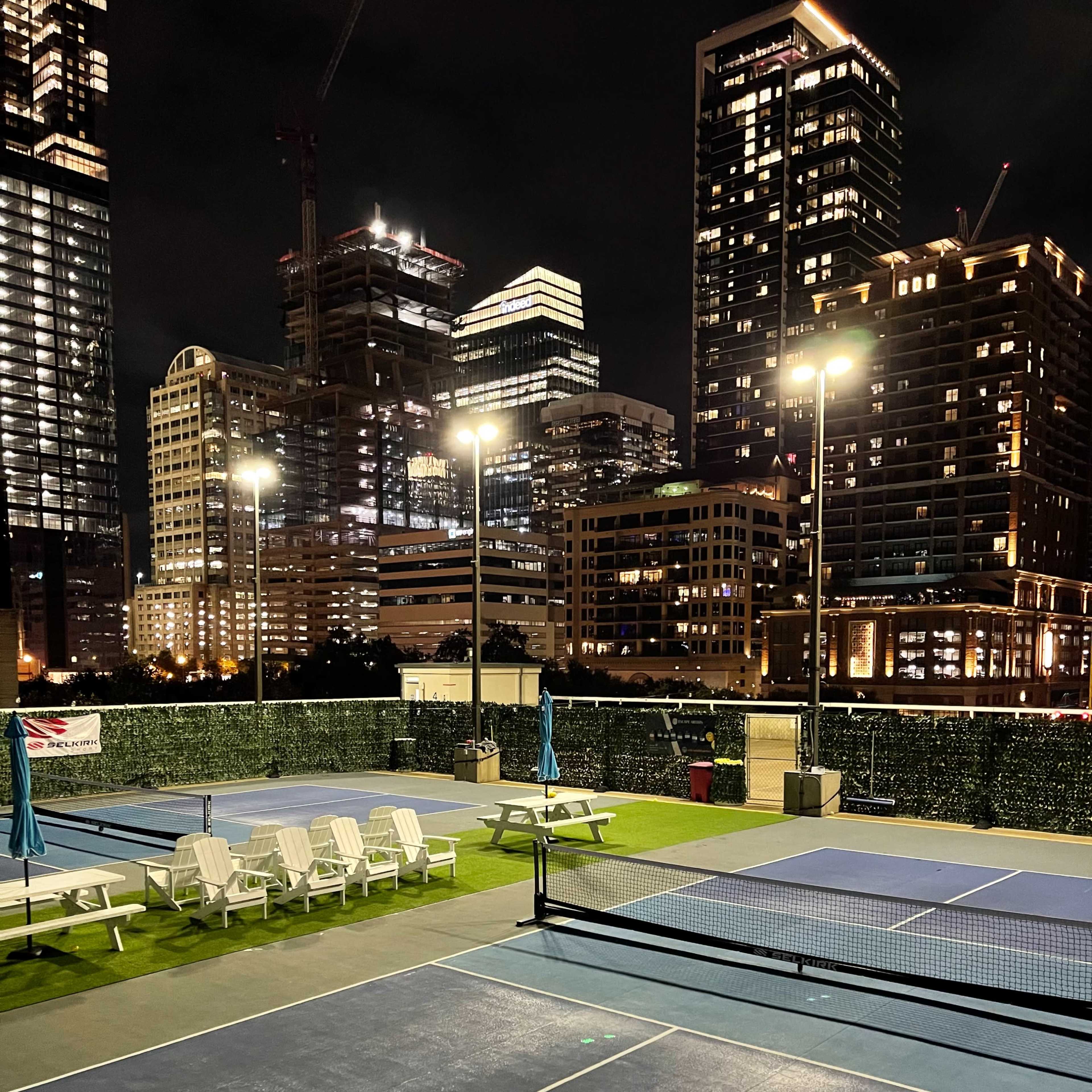 An empty tennis court at night, surrounded by lit buildings in an urban setting.