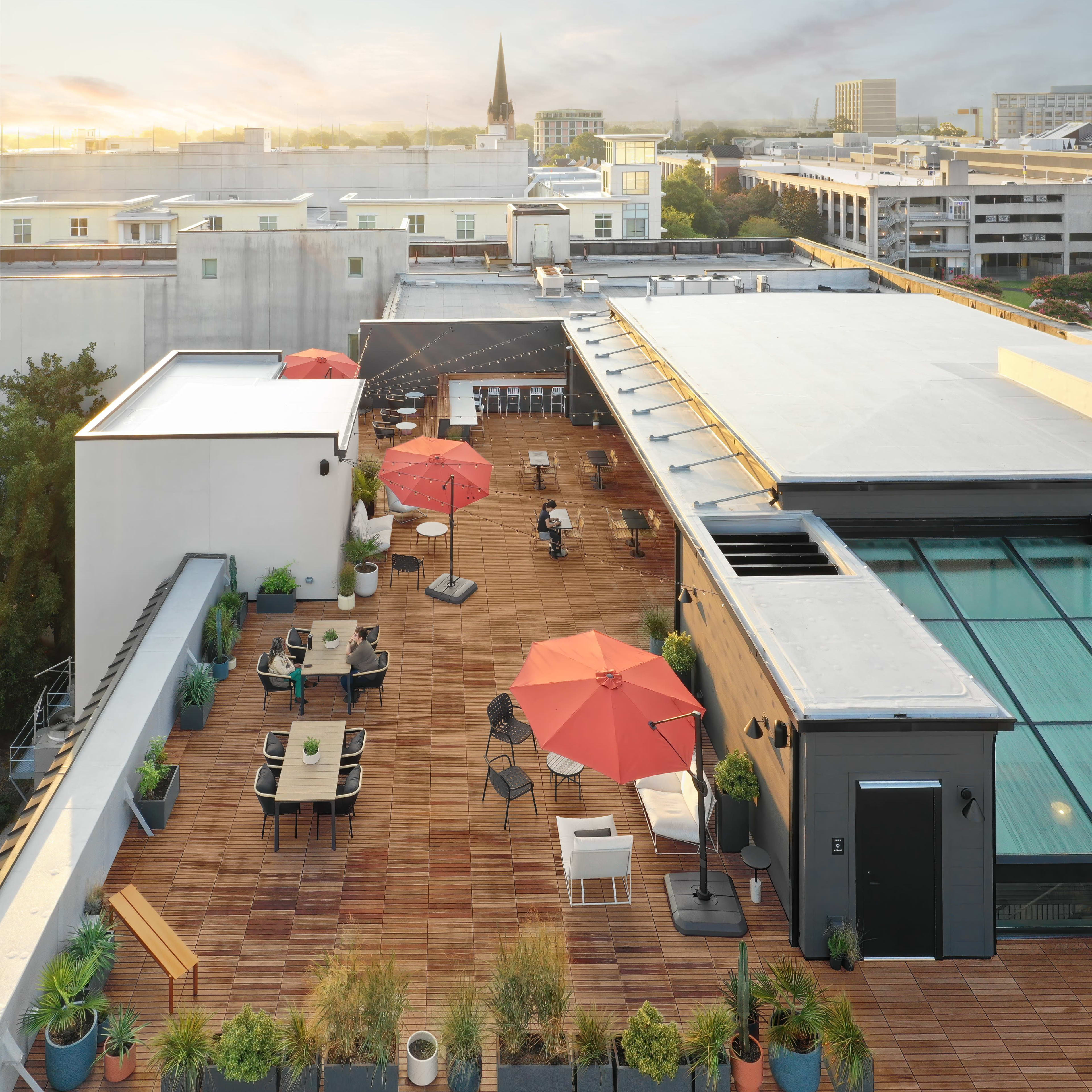 A spacious rooftop terrace with wooden flooring, outdoor furniture, and several red umbrellas, overlooking a cityscape at sunrise.