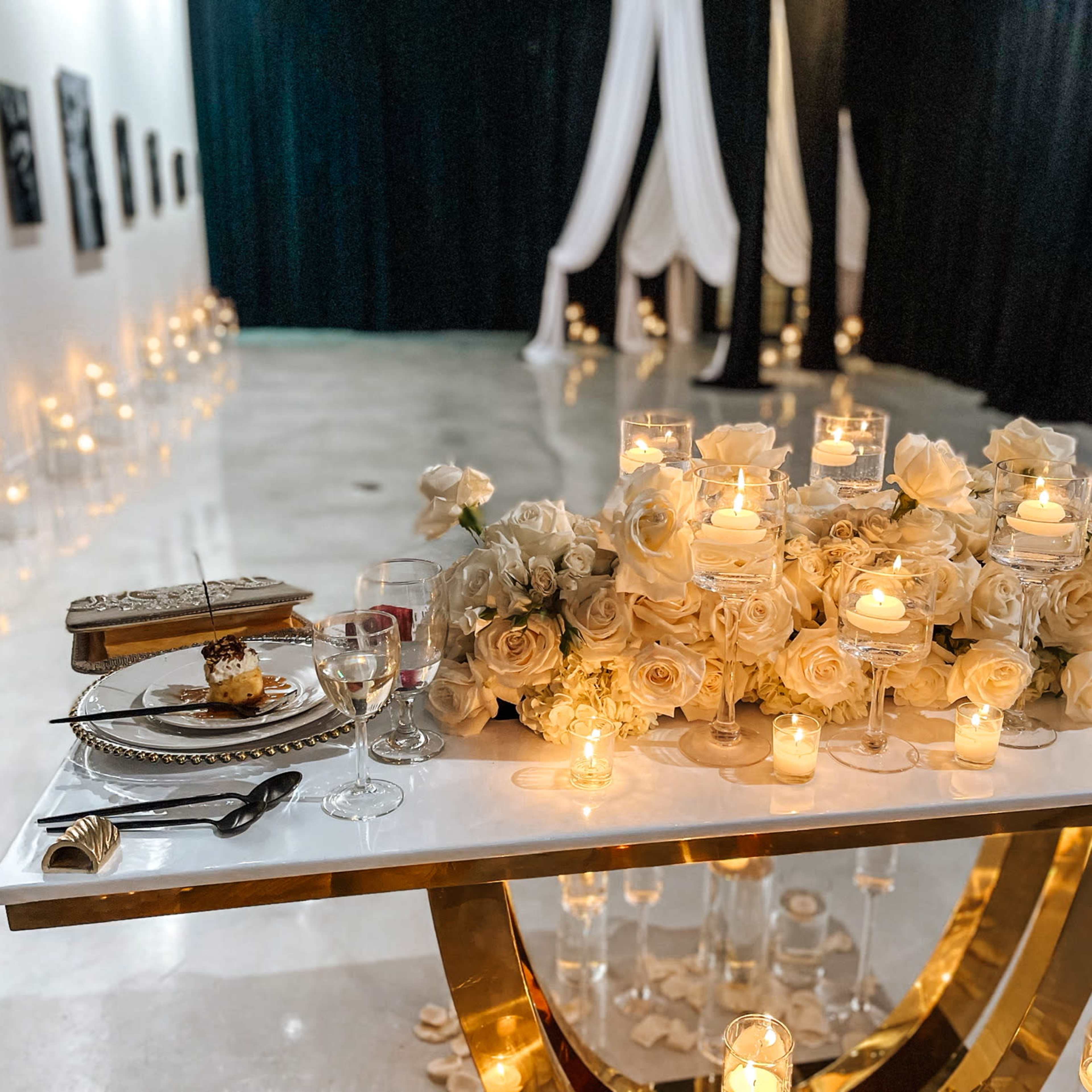 A beautifully arranged dining table is set with white roses and candles in a softly lit banquet hall, surrounded by elegant decorations and a backdrop of draped fabric.