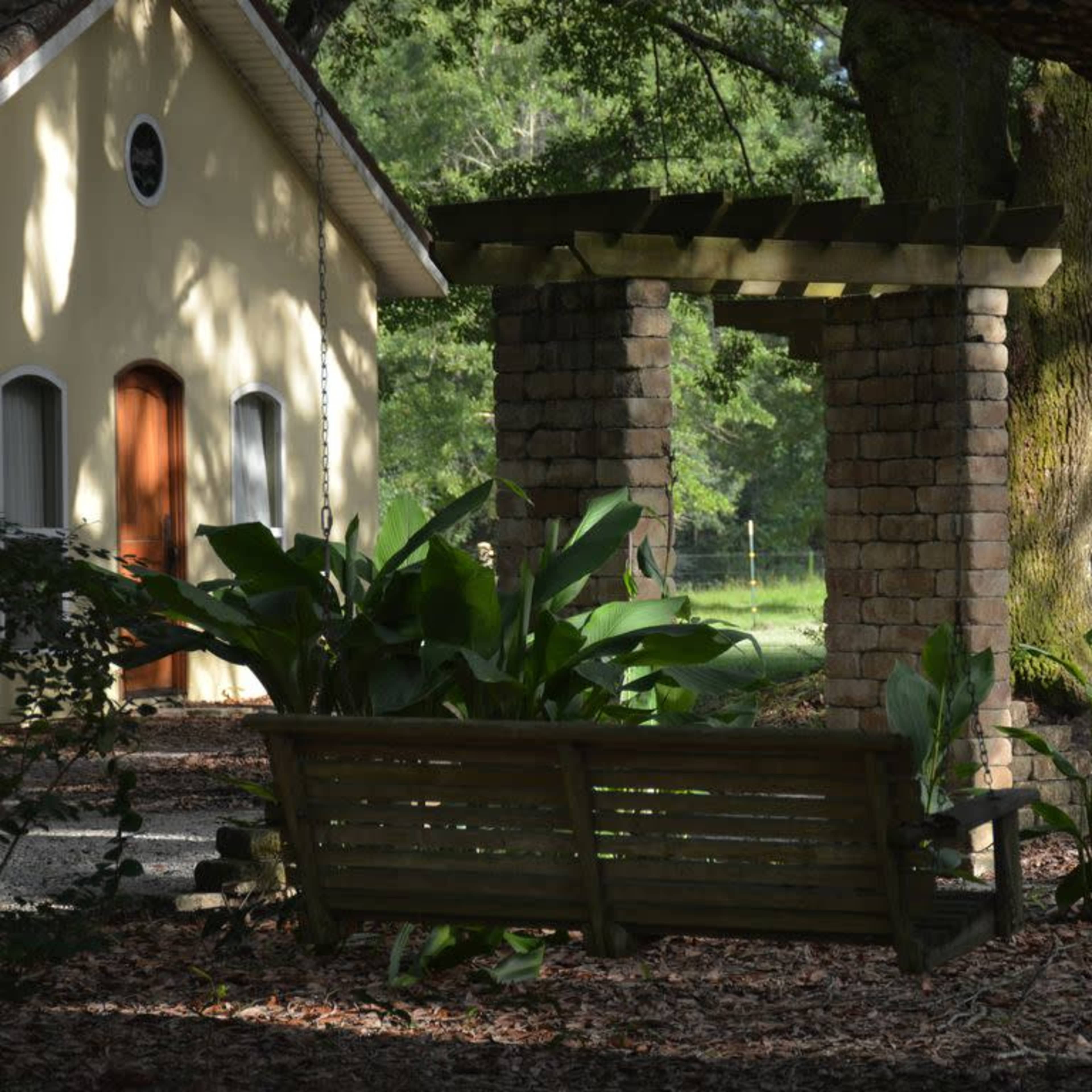 A wooden bench is positioned in front of a stone structure and a yellow building, surrounded by lush greenery and trees.