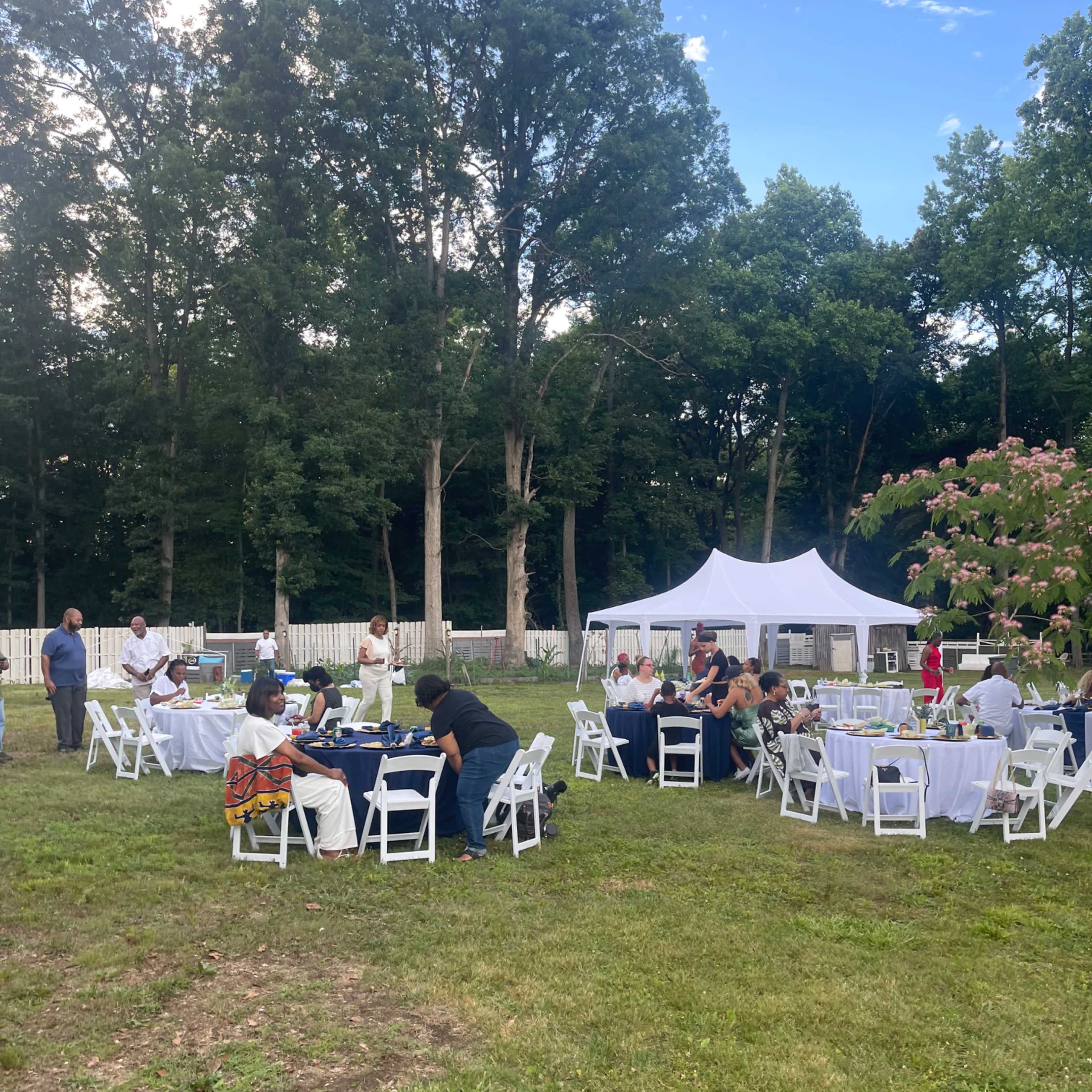 A group of people gather around tables under a tent in a grassy area surrounded by trees.