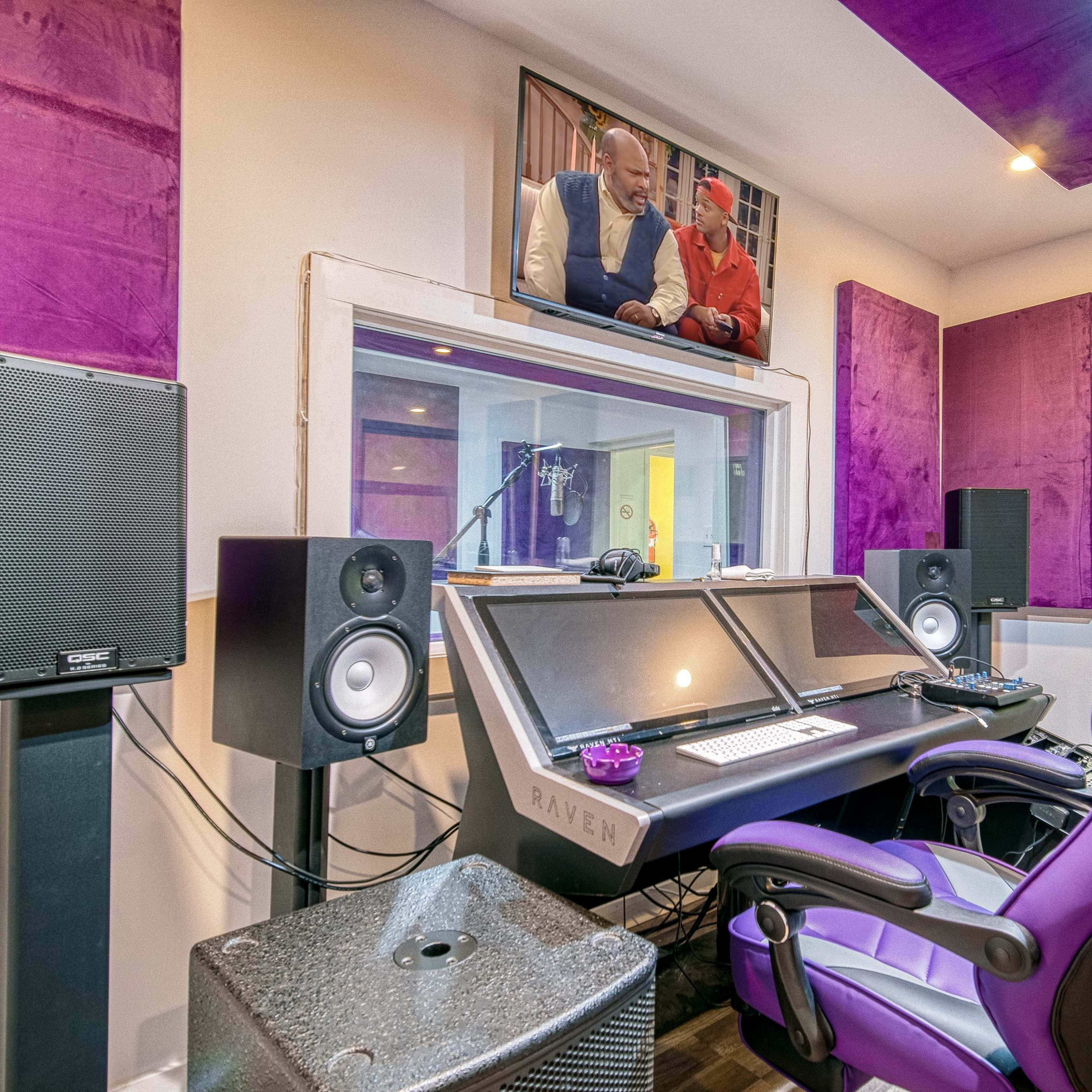A recording studio with a control desk featuring speakers, a computer monitor displaying a TV show, and purple soundproofing panels on the walls.