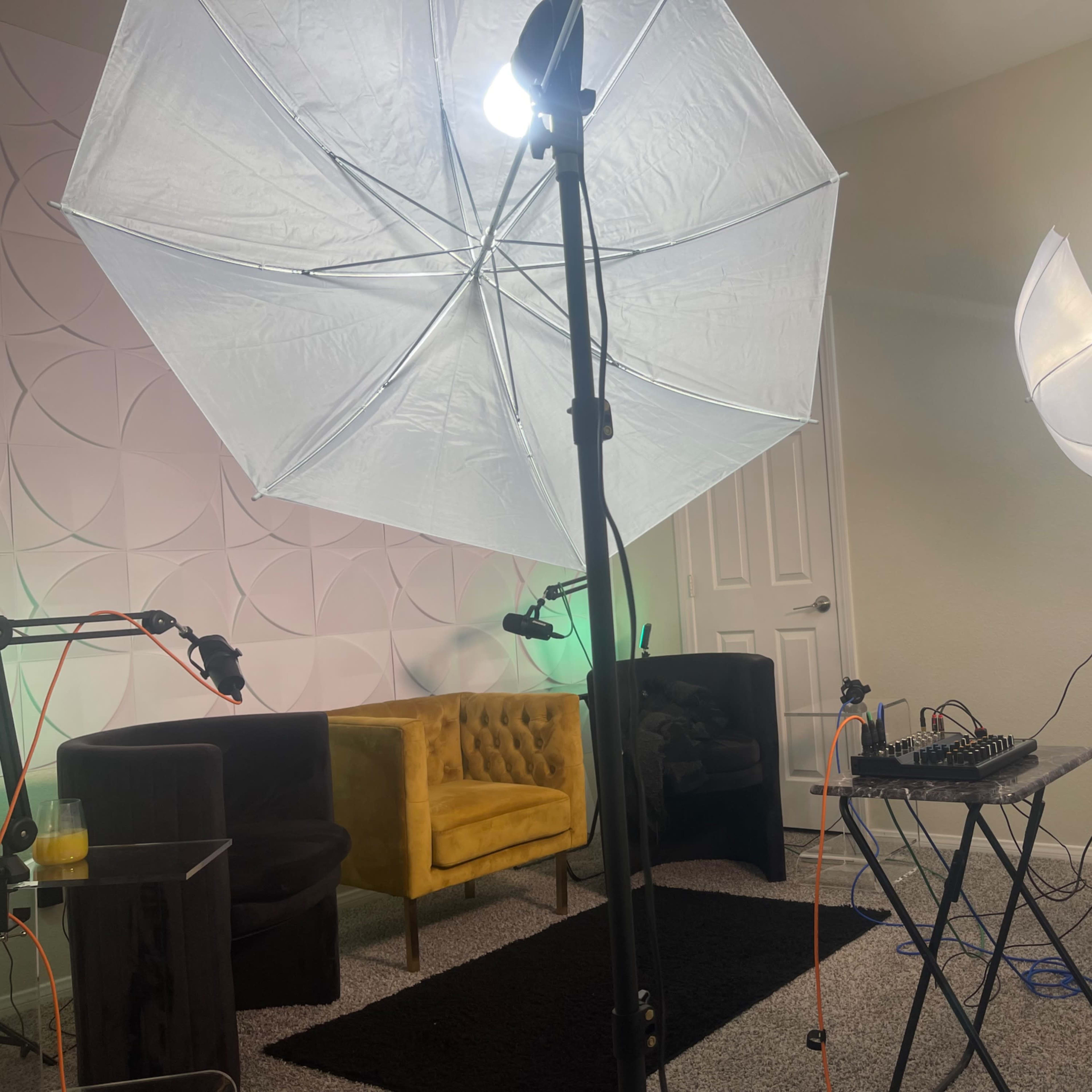 A brightly lit room features two large umbrella lights, a yellow upholstered chair, a black rug, and a table with audio equipment set up for recording.