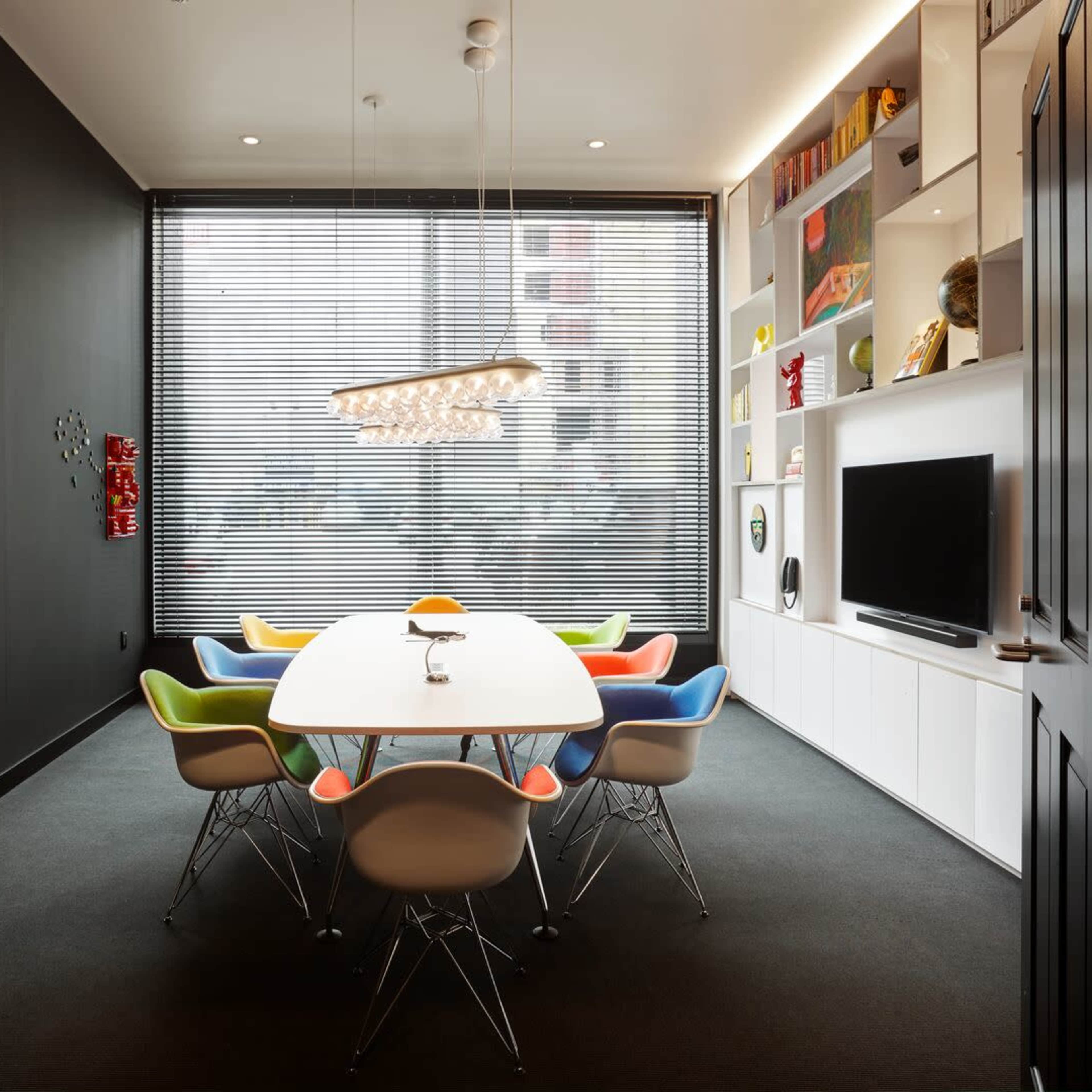 A modern conference room features a long white table surrounded by colorful chairs, with large windows that have slatted blinds.