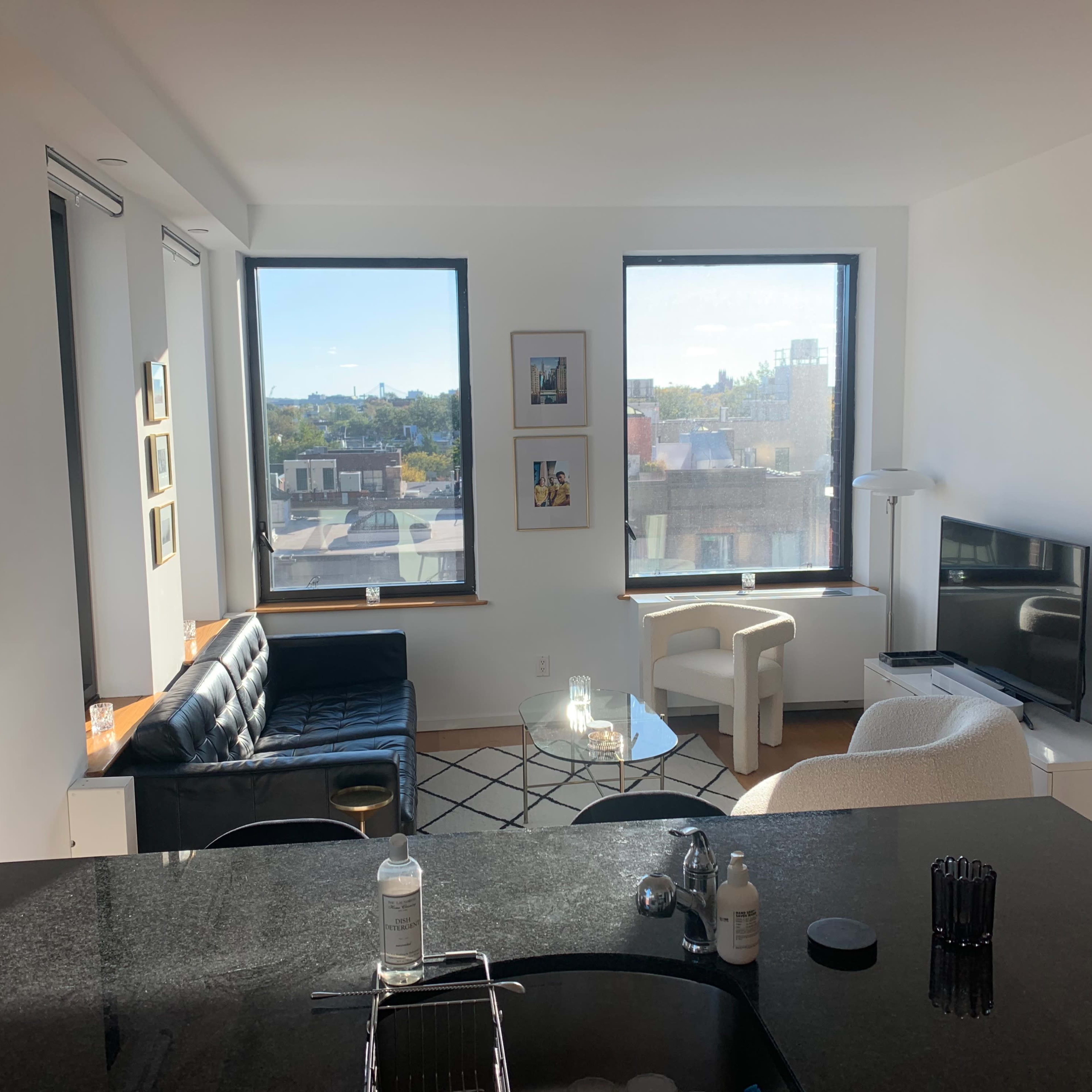 A modern living room features a black leather sofa, a white armchair, a glass coffee table, and a television, with large windows displaying a cityscape.