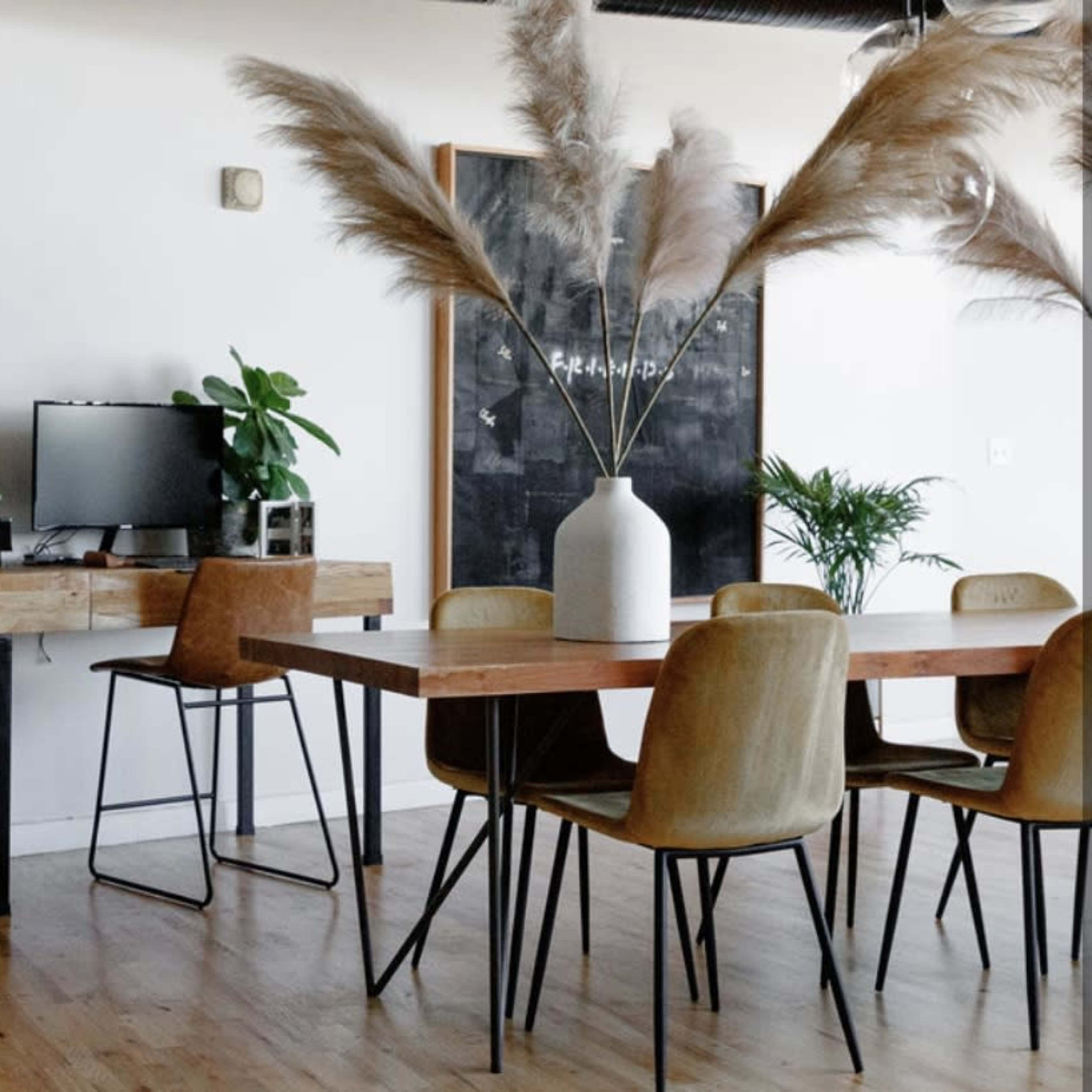 A modern office space with two computer desks facing a dining table surrounded by chairs, all complemented by decorative plants and tall pampas grass in a vase.