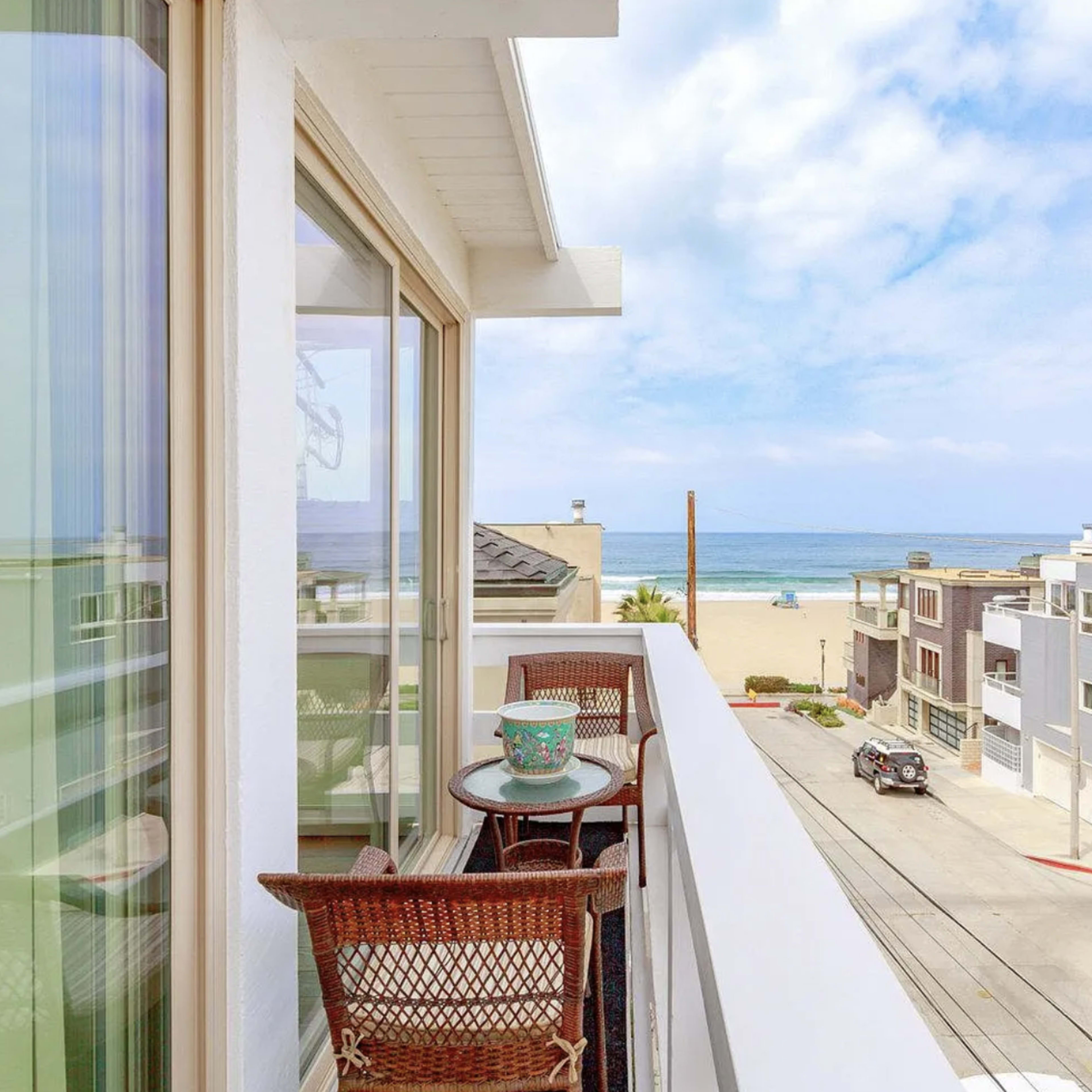 A balcony with wicker chairs overlooks a sandy beach and ocean, framed by nearby modern buildings.