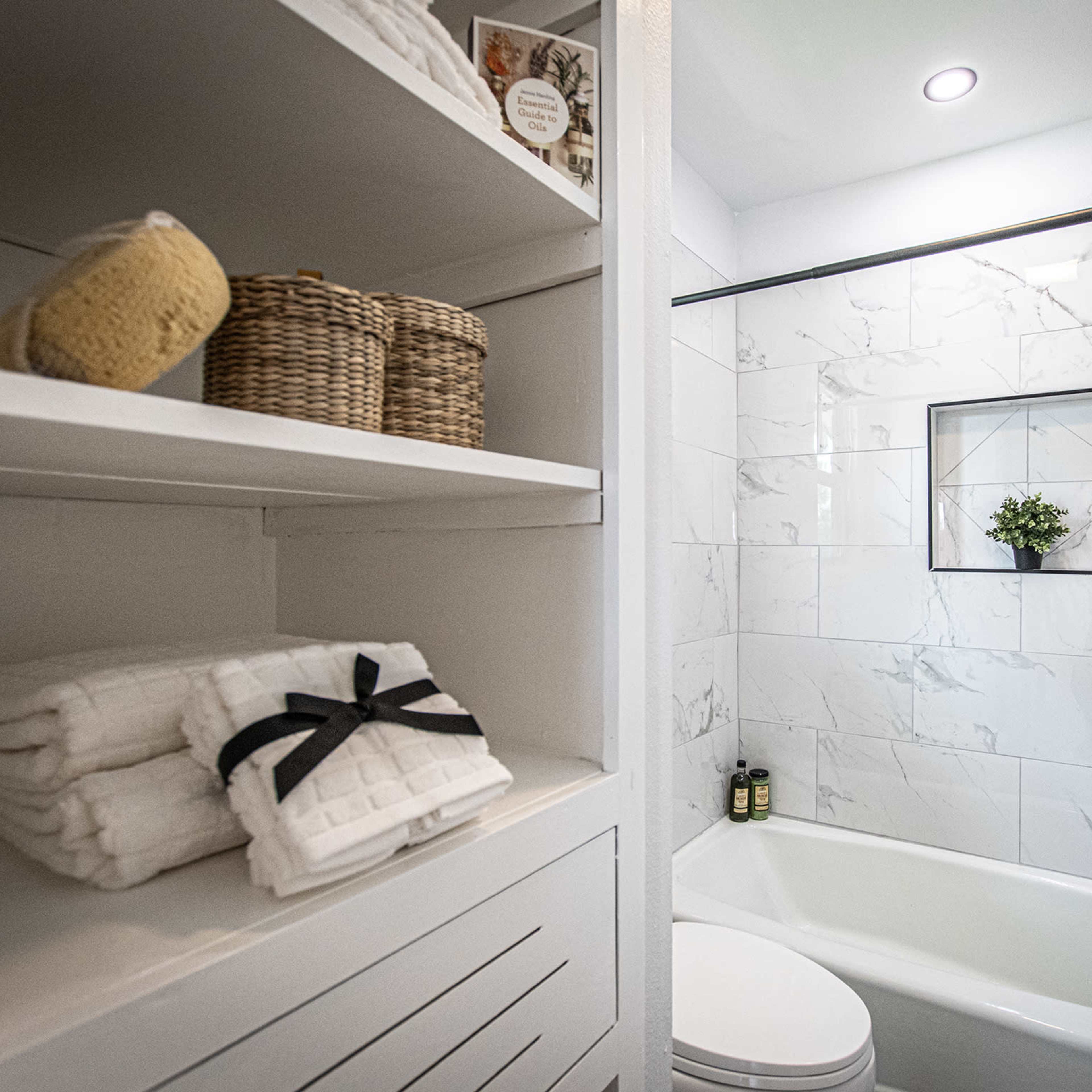 A modern bathroom with white tiles, a bathtub, a storage shelf displaying neatly folded towels and baskets, and a small plant on the wall.