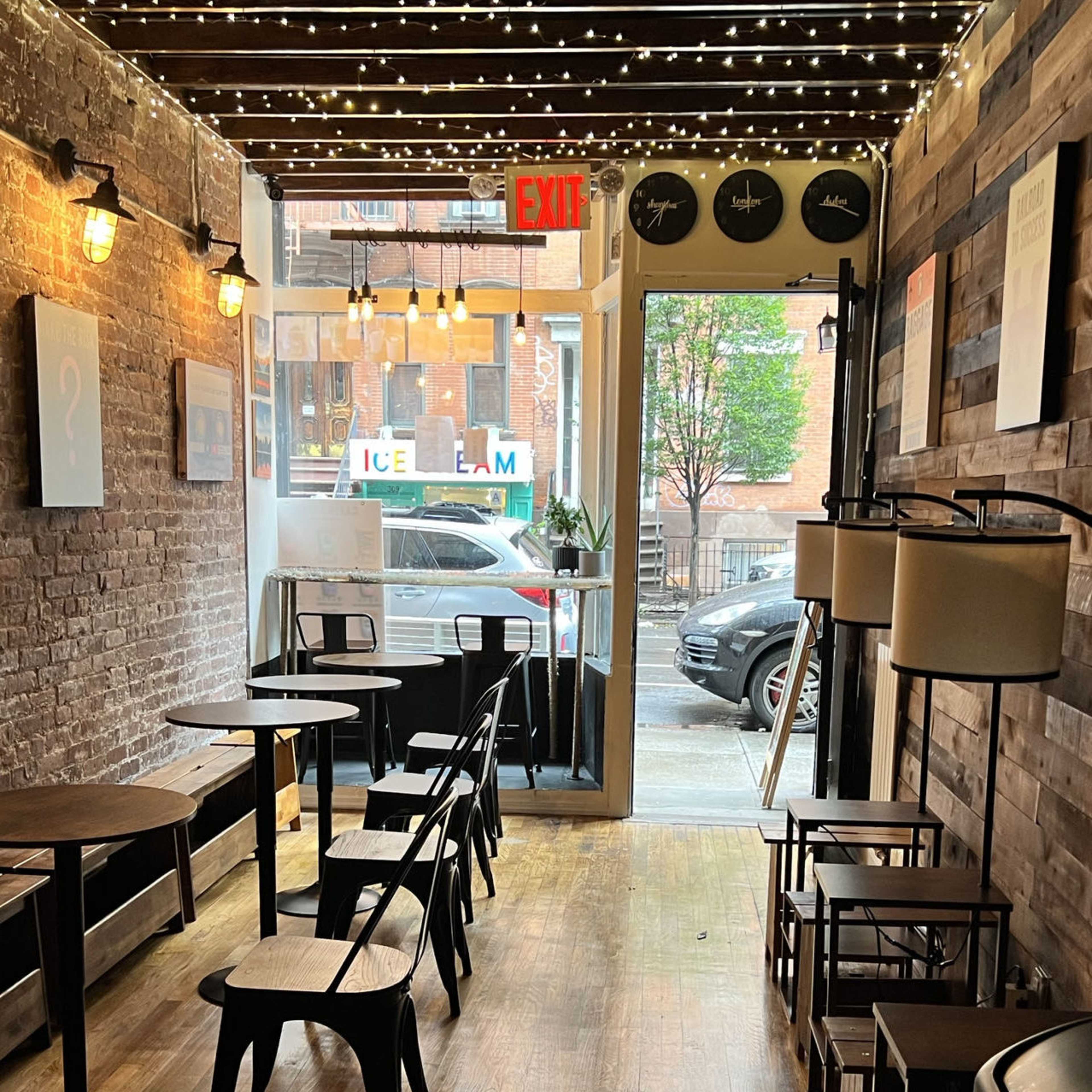 A narrow café with wooden flooring and exposed brick walls, featuring several tables and chairs arranged along the length of the space, leading to a doorway with an exit sign.