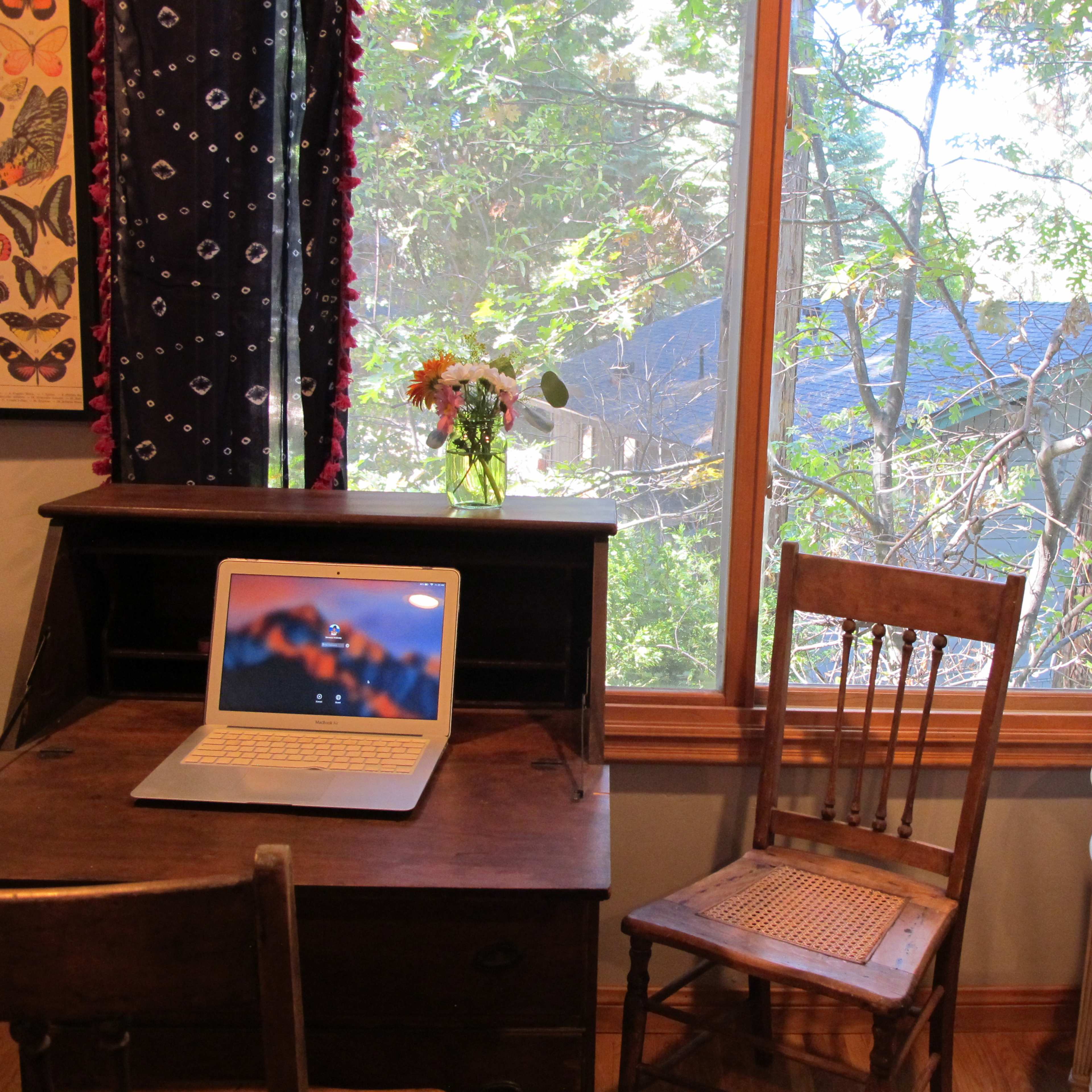 A wooden desk with a laptop and a chair is positioned near a window overlooking trees and a glimpse of a roof outside.