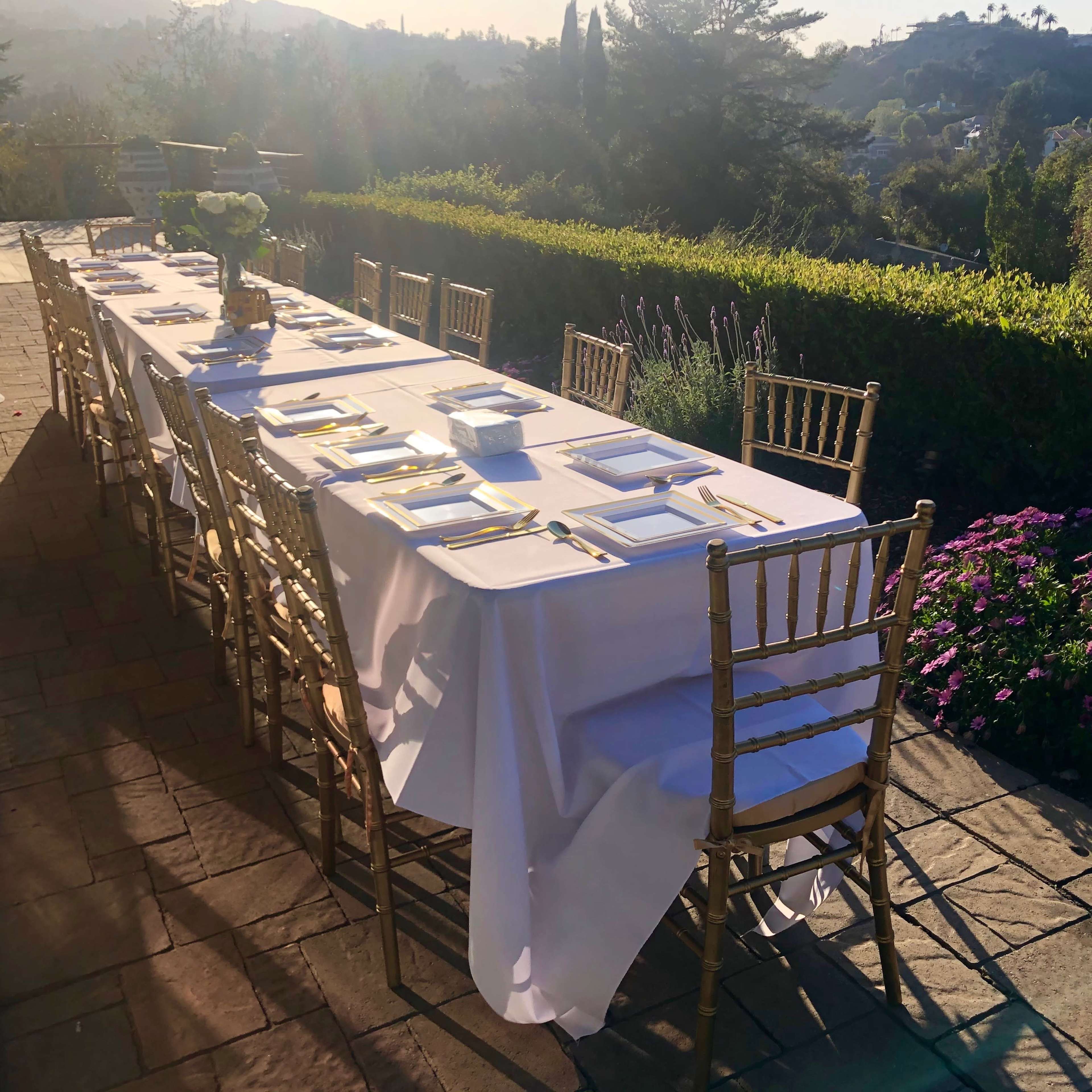 A long dining table with white tablecloth and gold chairs is set outdoors on a stone patio, surrounded by greenery and flowers.