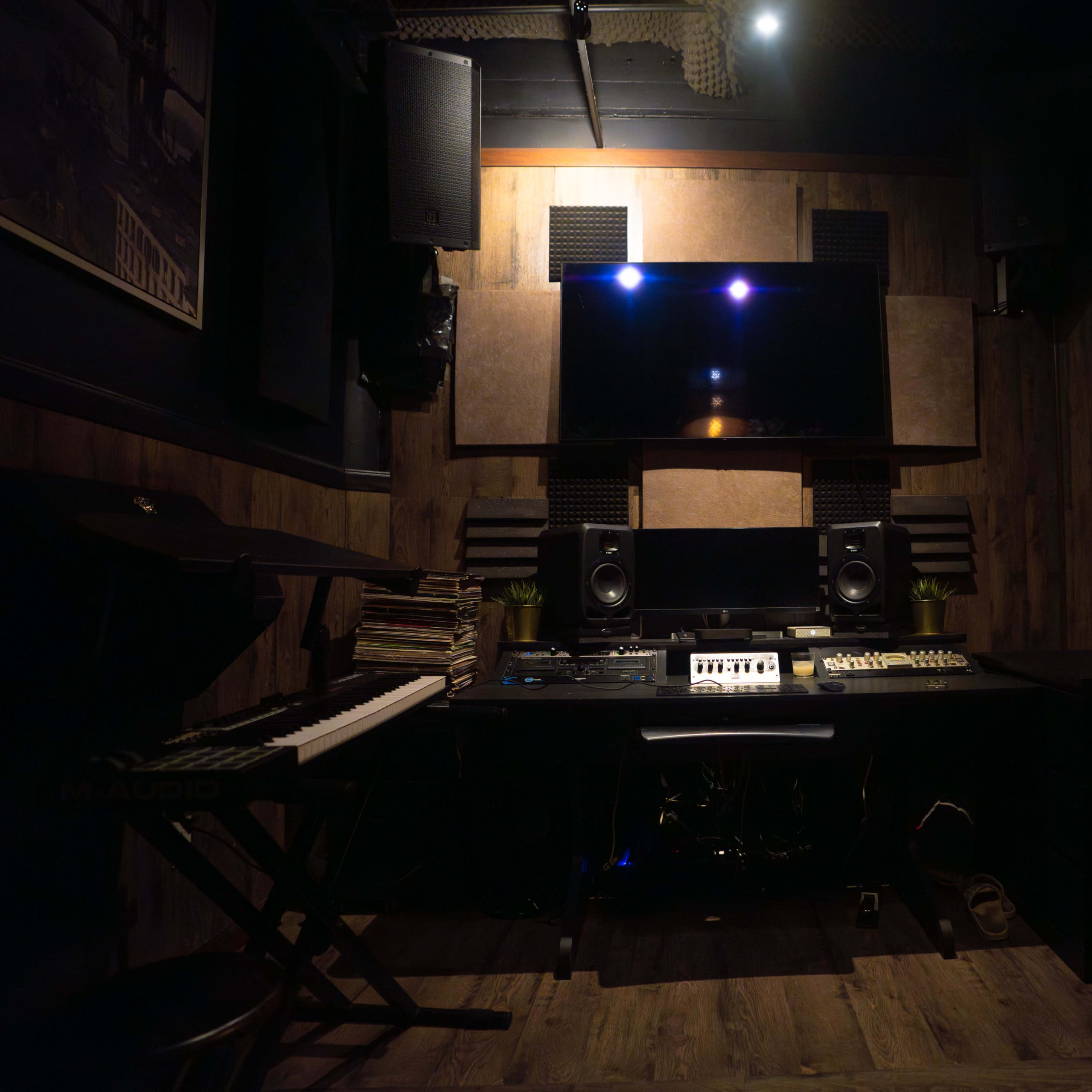 A music studio features a wooden interior, a keyboard on the left, speakers mounted on the walls, and a central console with audio equipment.