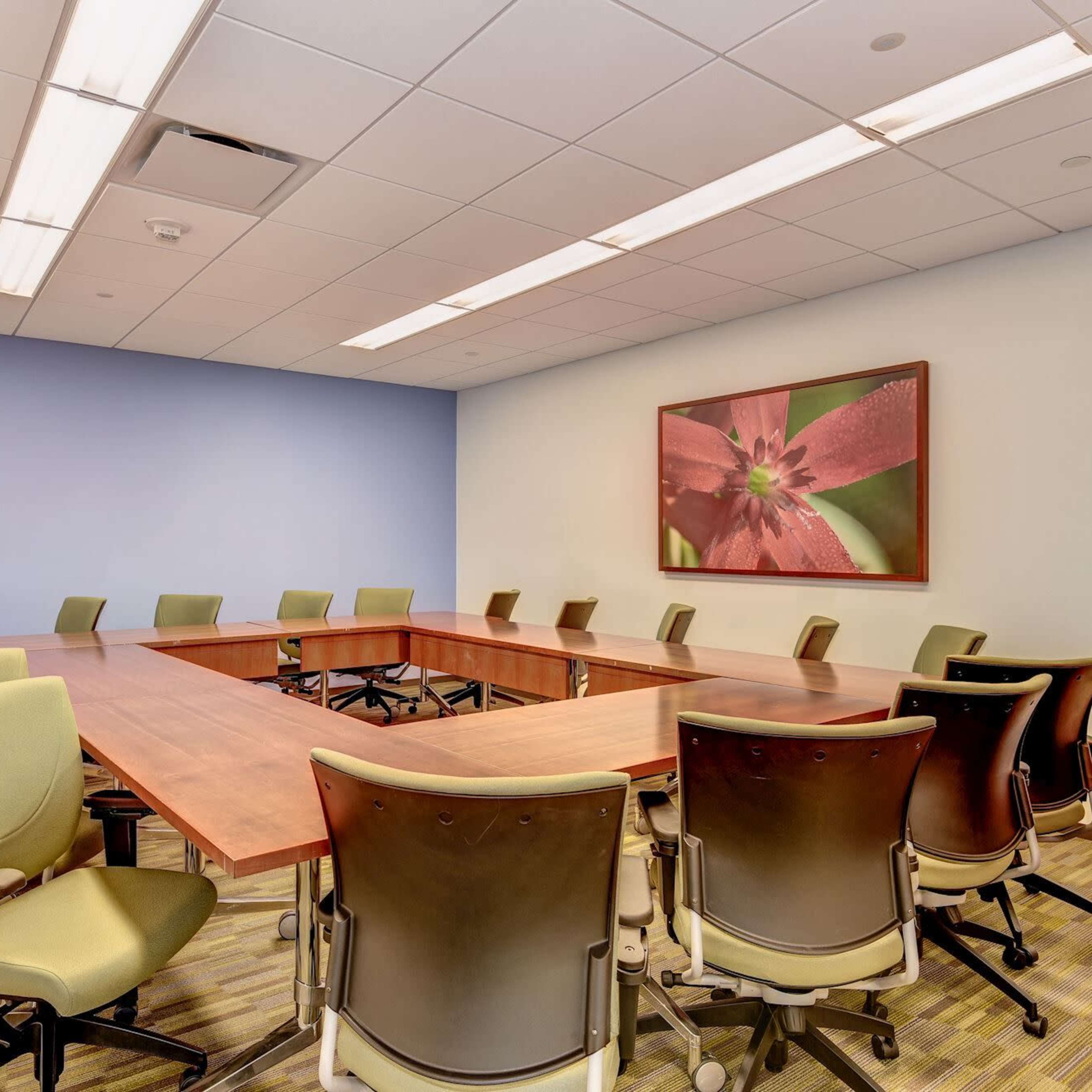 A conference room features a large wooden table surrounded by ergonomic chairs, with a floral artwork on the wall and blue accents.