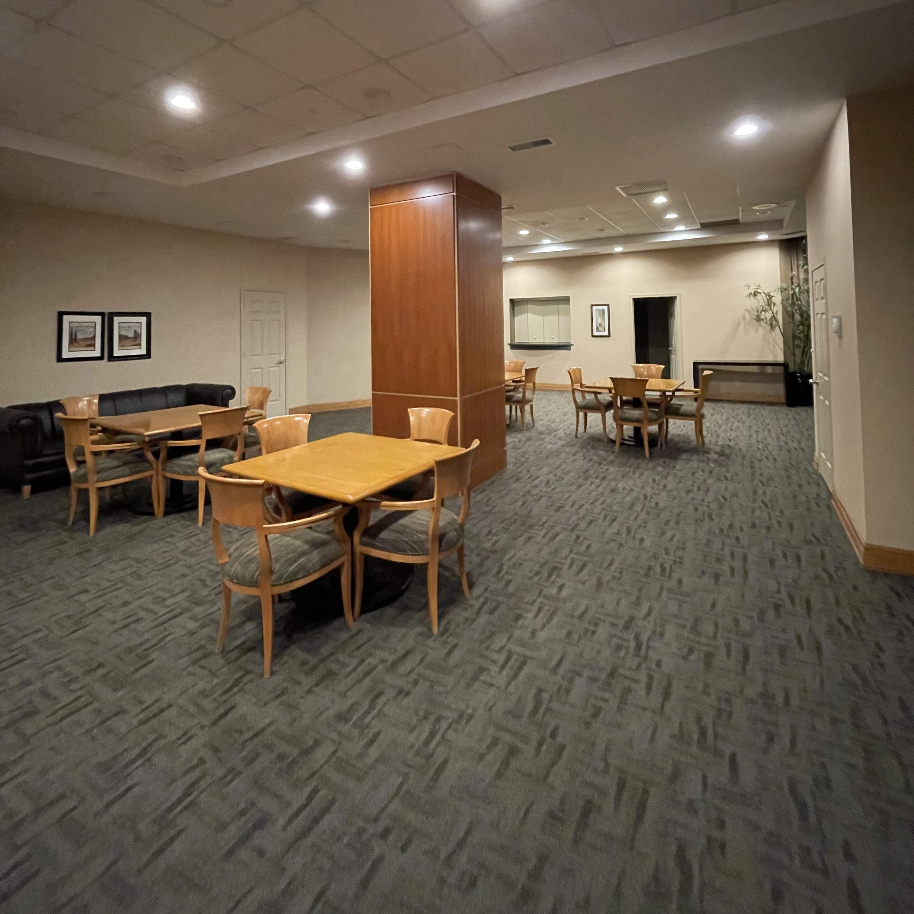 A spacious lounge area features wooden tables and chairs arranged around a central wooden column, with a carpeted floor and soft overhead lighting.