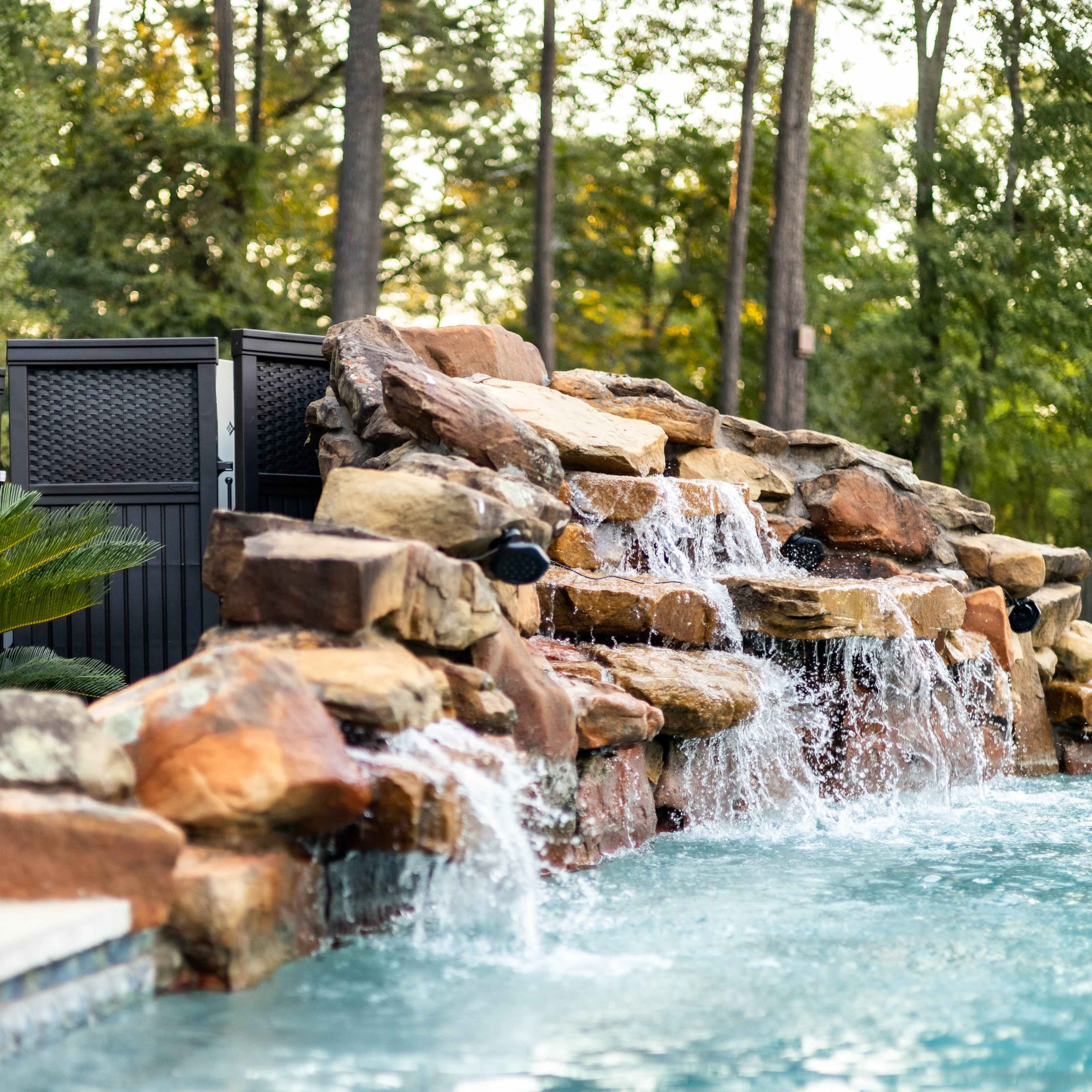 A stone waterfall cascades into a blue pool surrounded by greenery and a privacy screen in a wooded backyard.