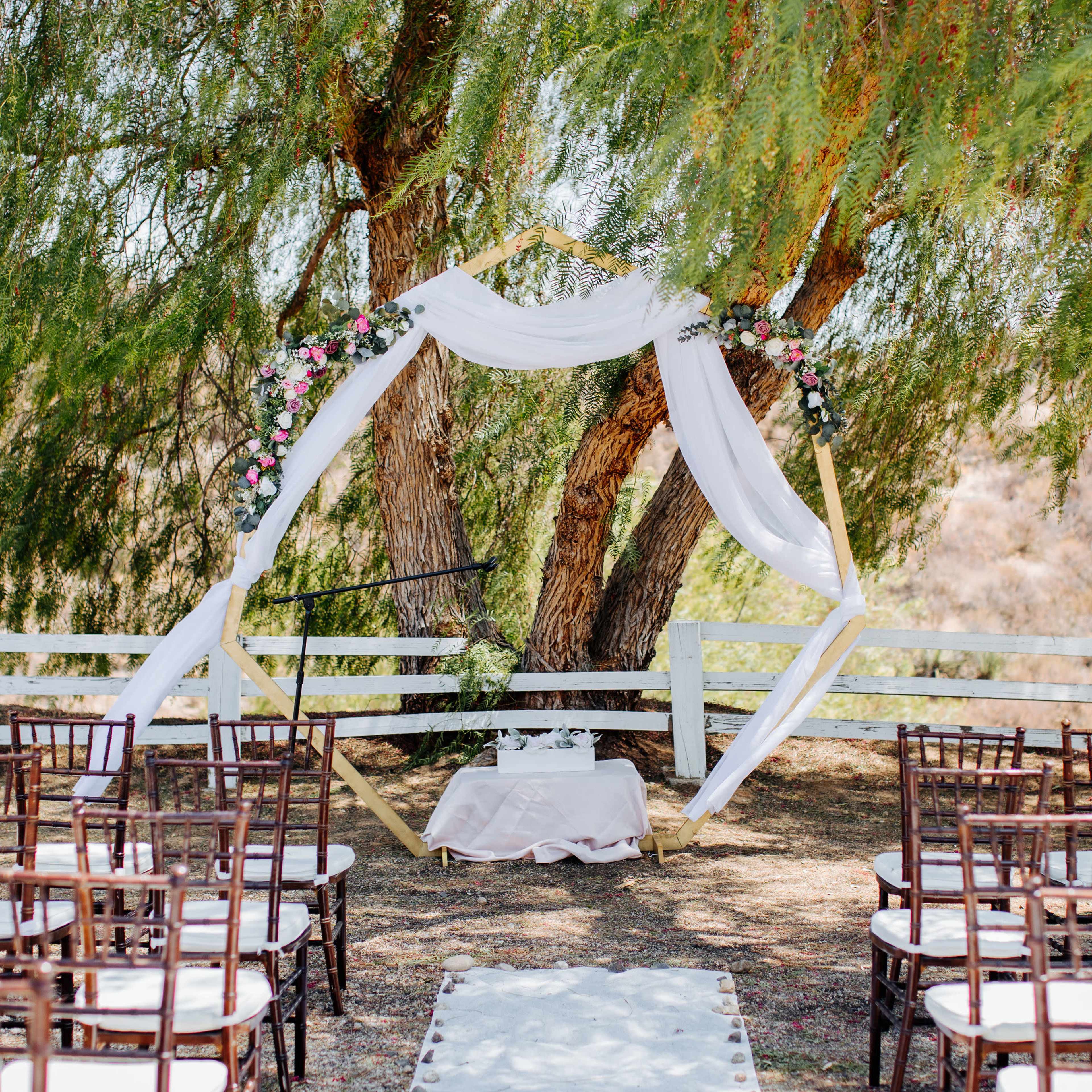 A wedding ceremony setup features an elegant altar draped in white fabric under two large trees, with rows of chairs arranged in front.