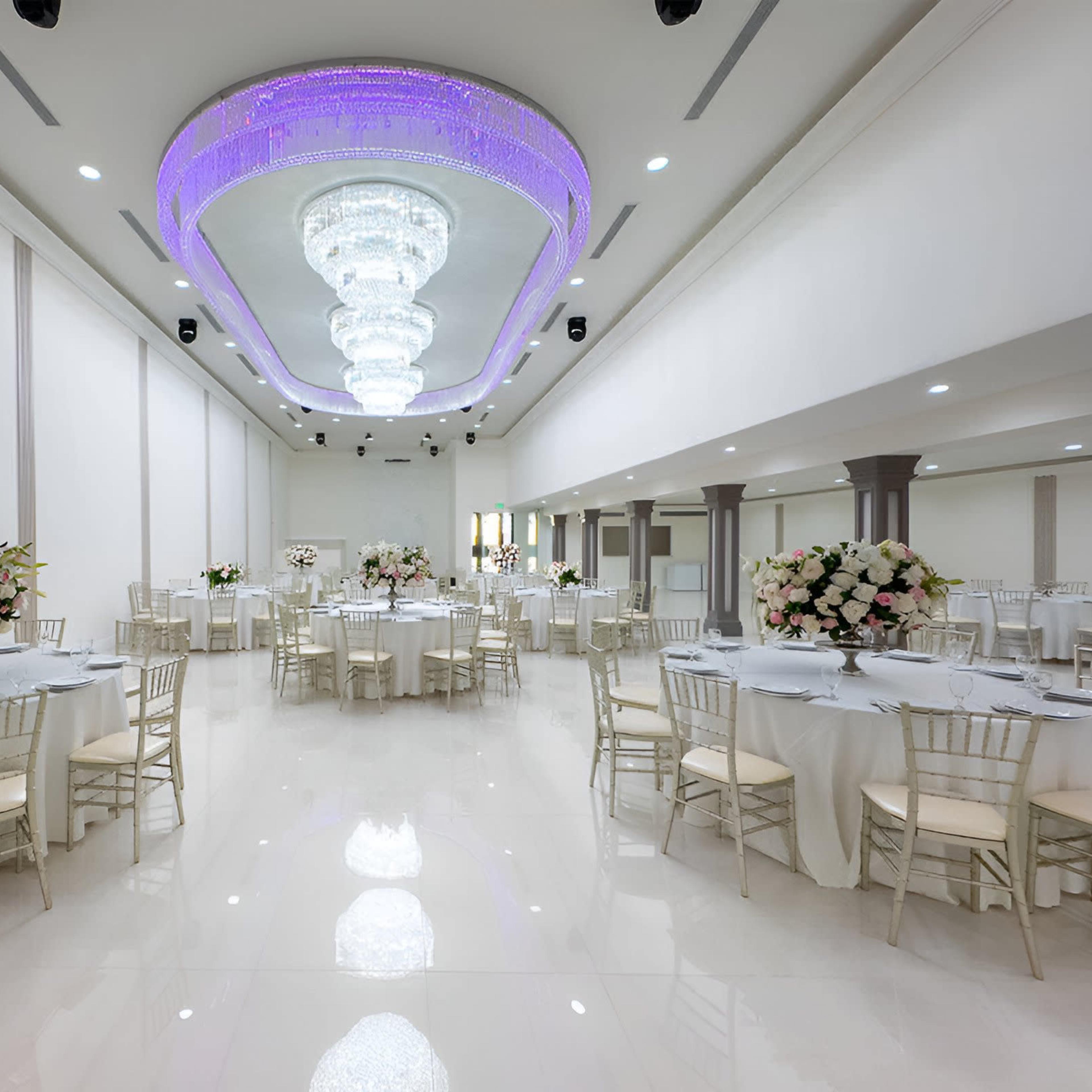 A spacious banquet hall features round tables adorned with floral centerpieces and a large crystal chandelier illuminated by purple lighting.