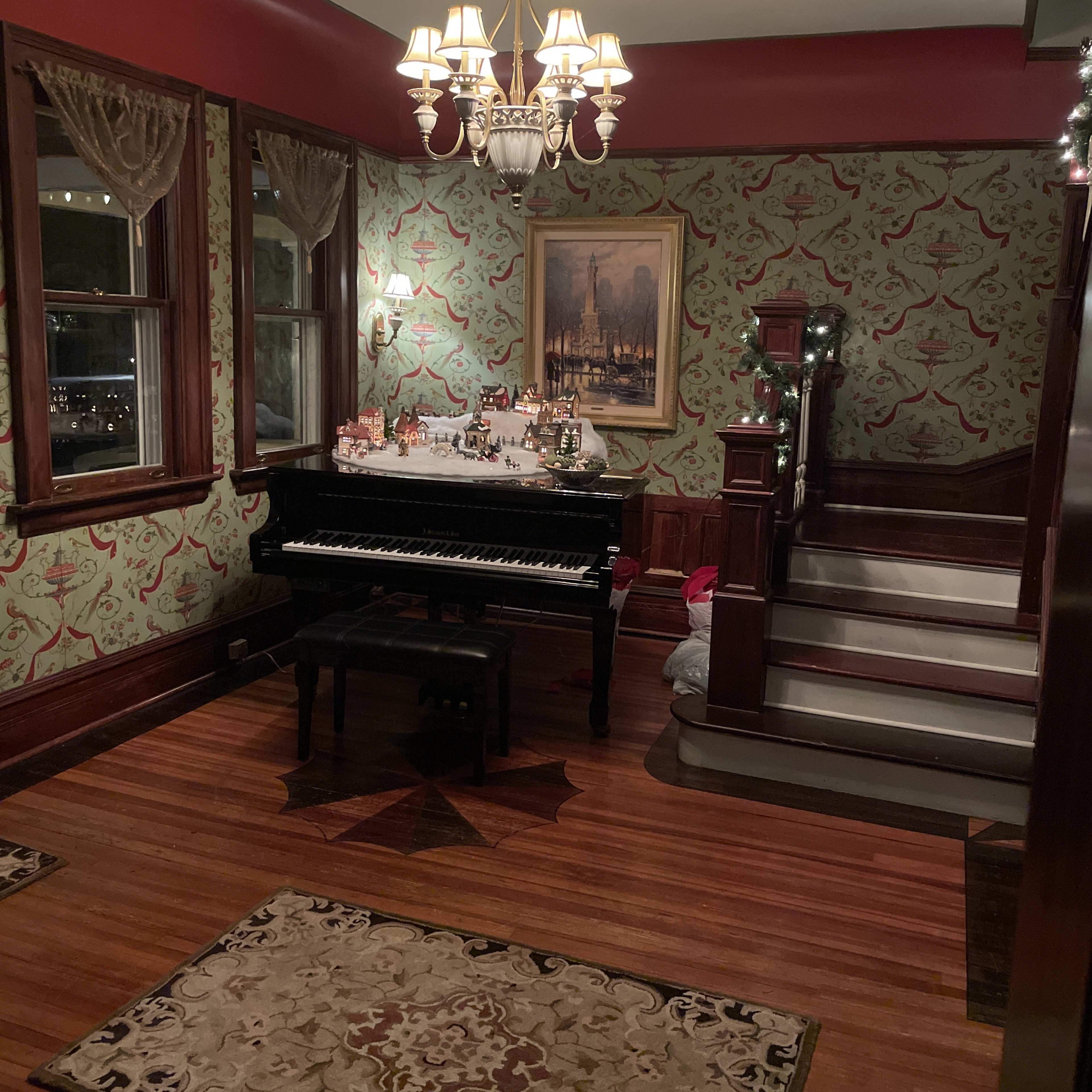 A warmly lit room featuring a black piano, a decorative chandelier, and ornate wallpaper, with a staircase visible in the background.