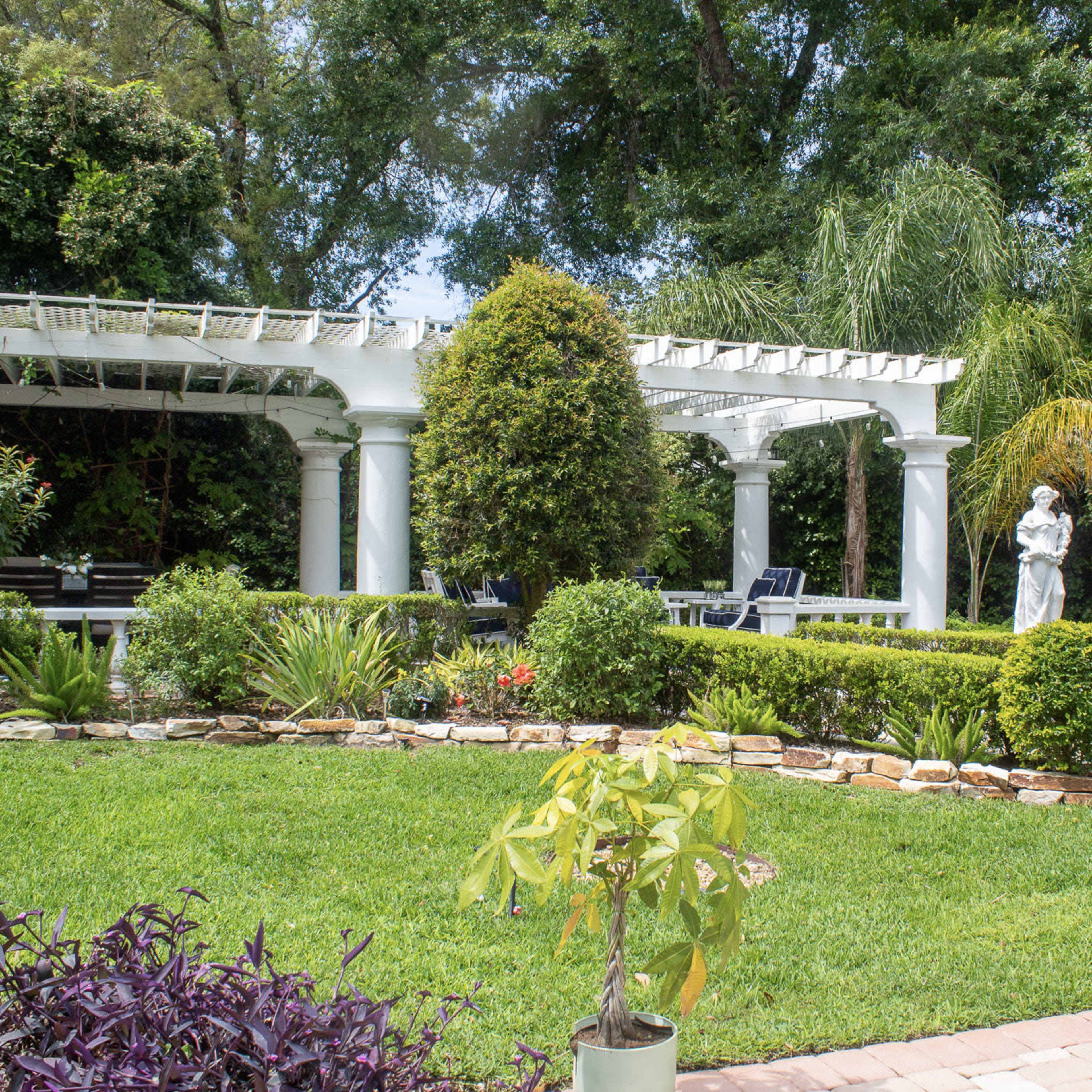 A landscaped garden featuring a stone pathway, greenery, a white pergola, and a statue amidst various plants.