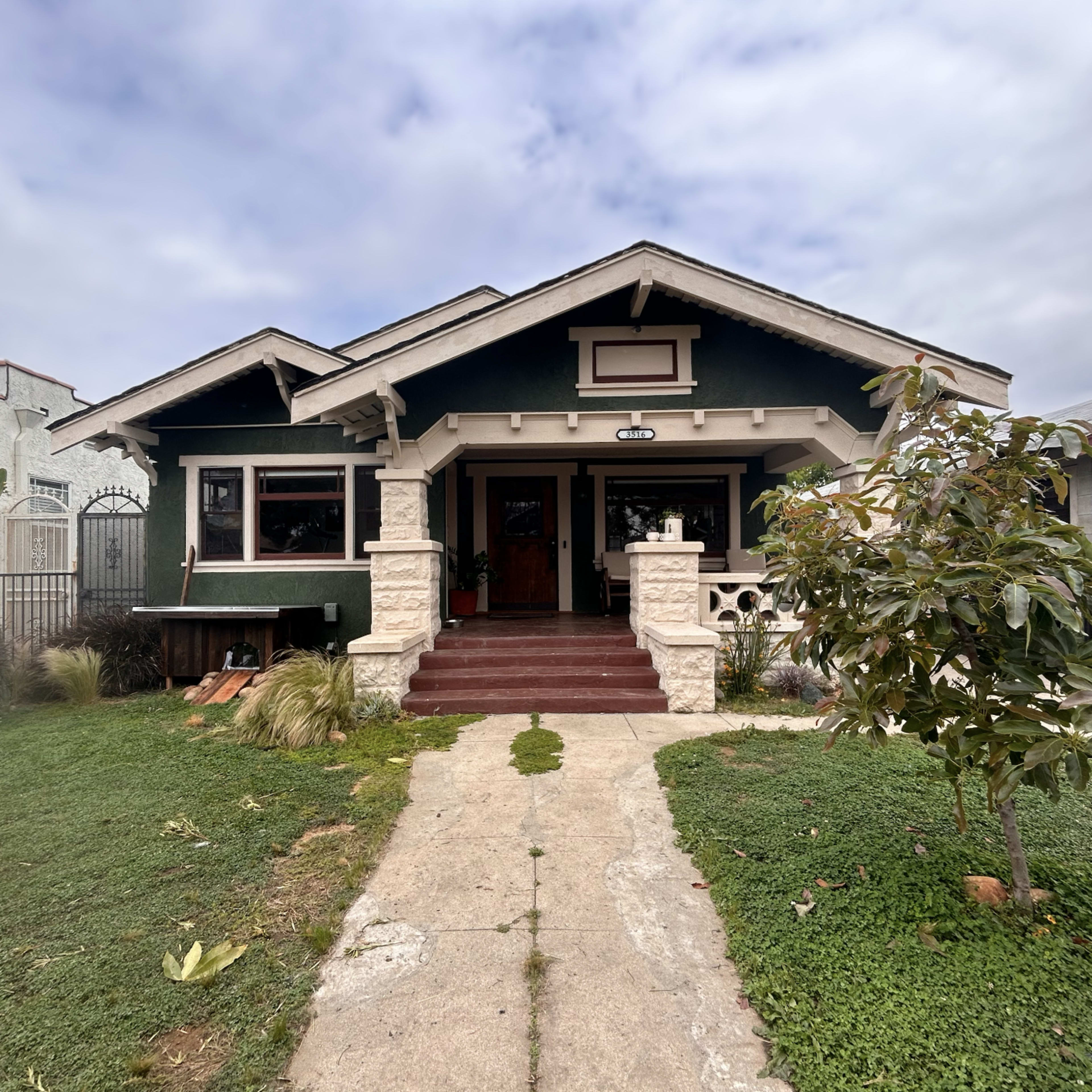 A green bungalow-style house with a porch, surrounded by grass and a small tree in the front yard.