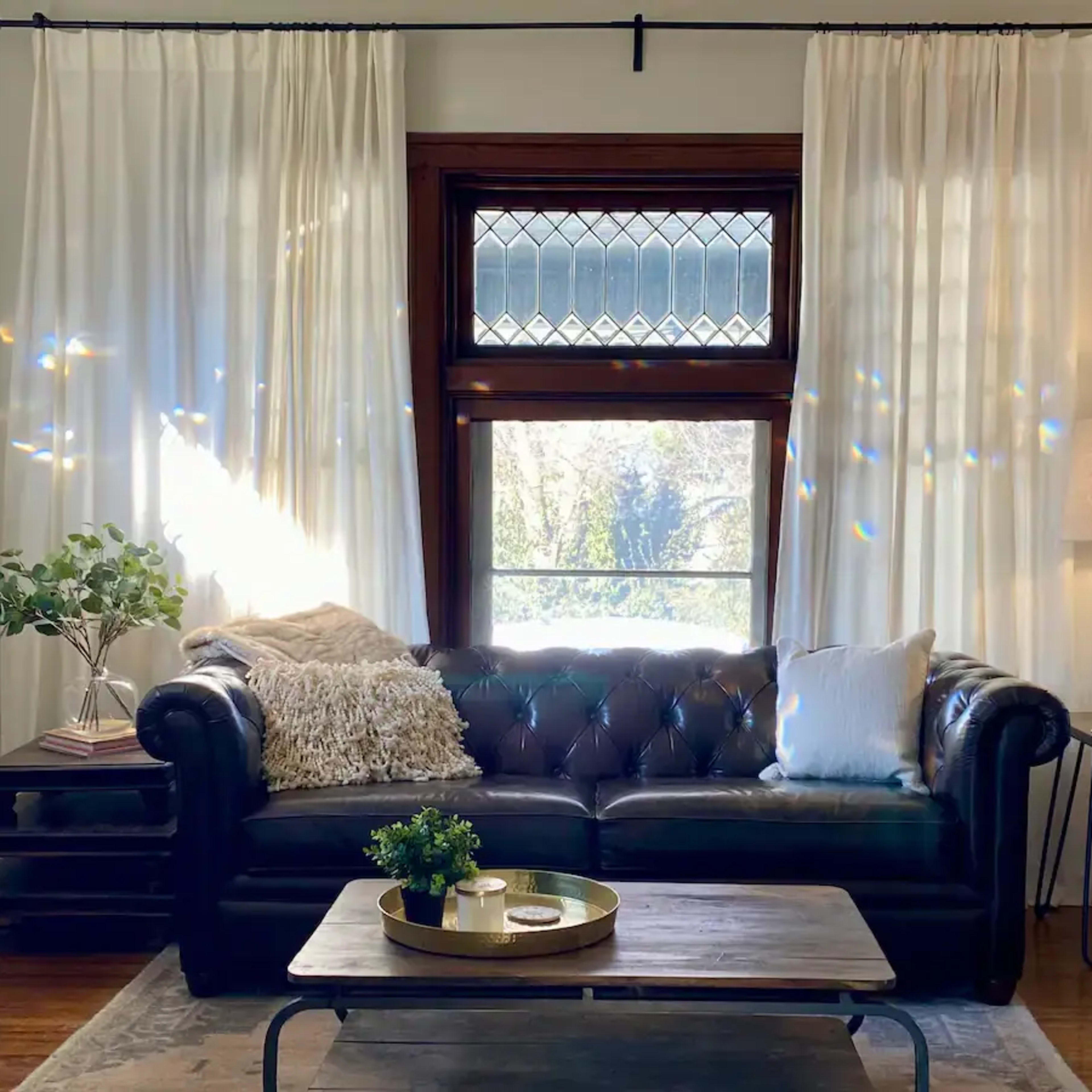 A leather sofa is positioned in a living room, accompanied by a coffee table and a floor lamp, with sheer curtains framing a window that lets in natural light.