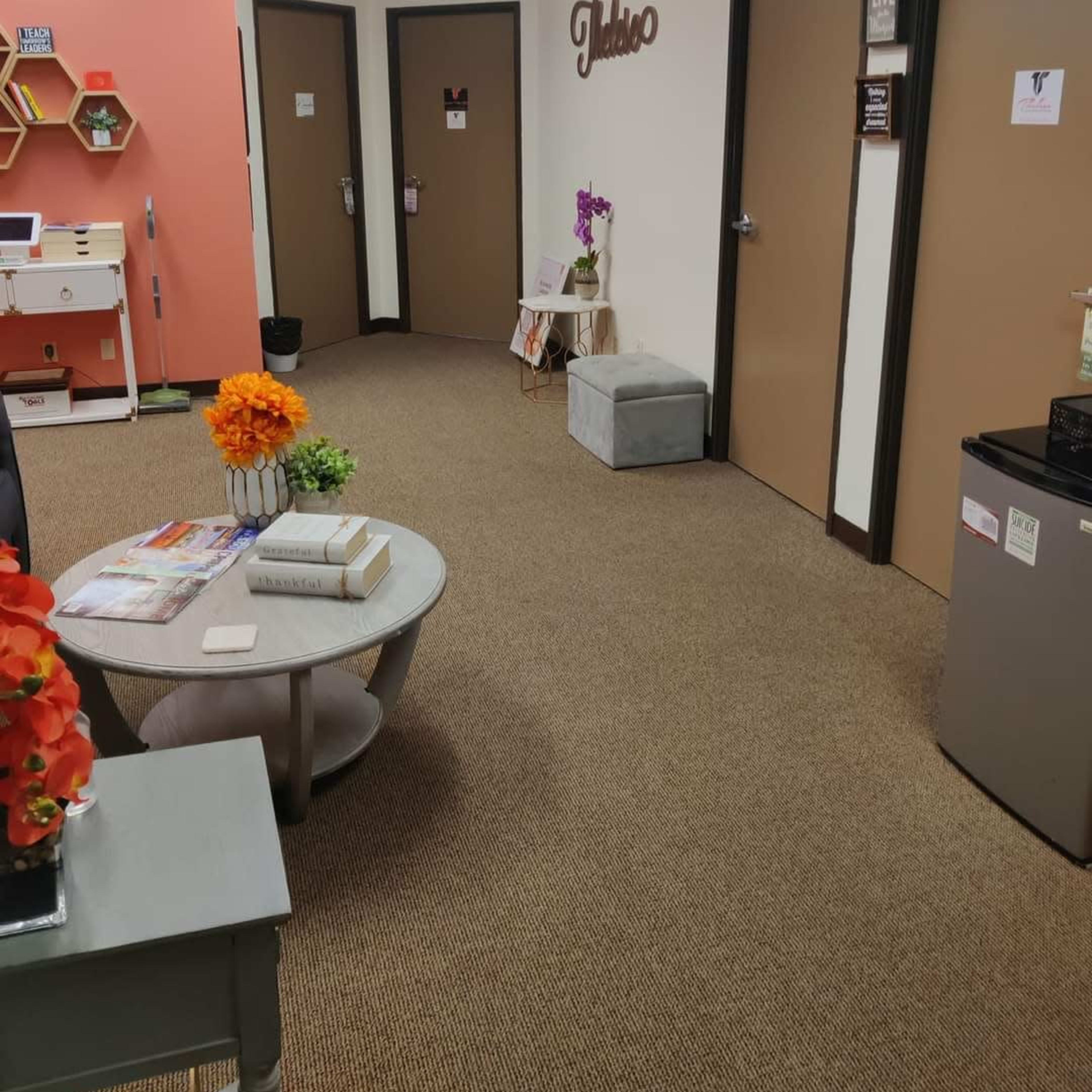 A hallway with beige carpet, featuring three closed doors, a small decorative table with a flower arrangement, and a seating area with a round table and magazines.