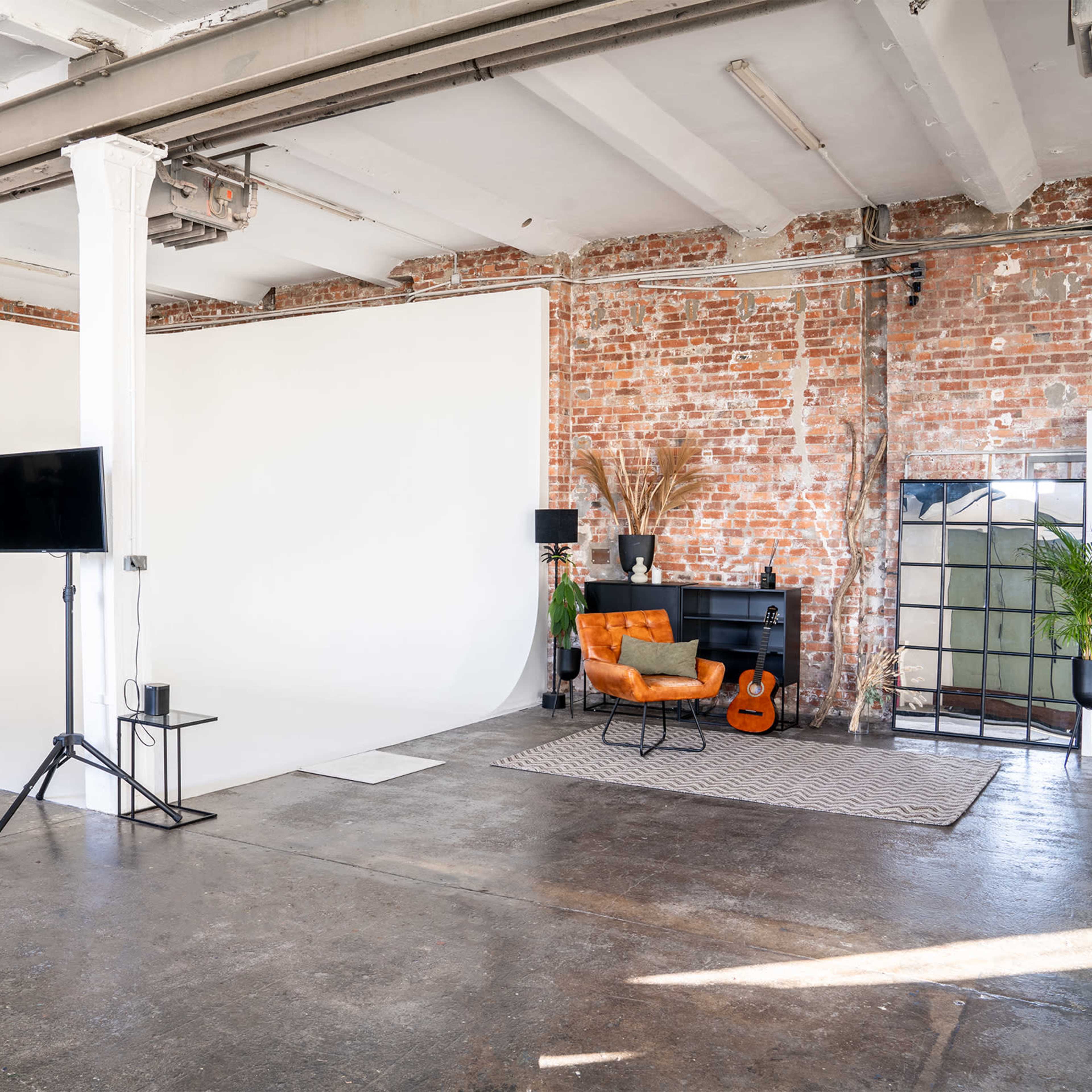 A spacious studio with a white backdrop, exposed brick walls, and a cozy seating area featuring an orange chair, a guitar, and potted plants.