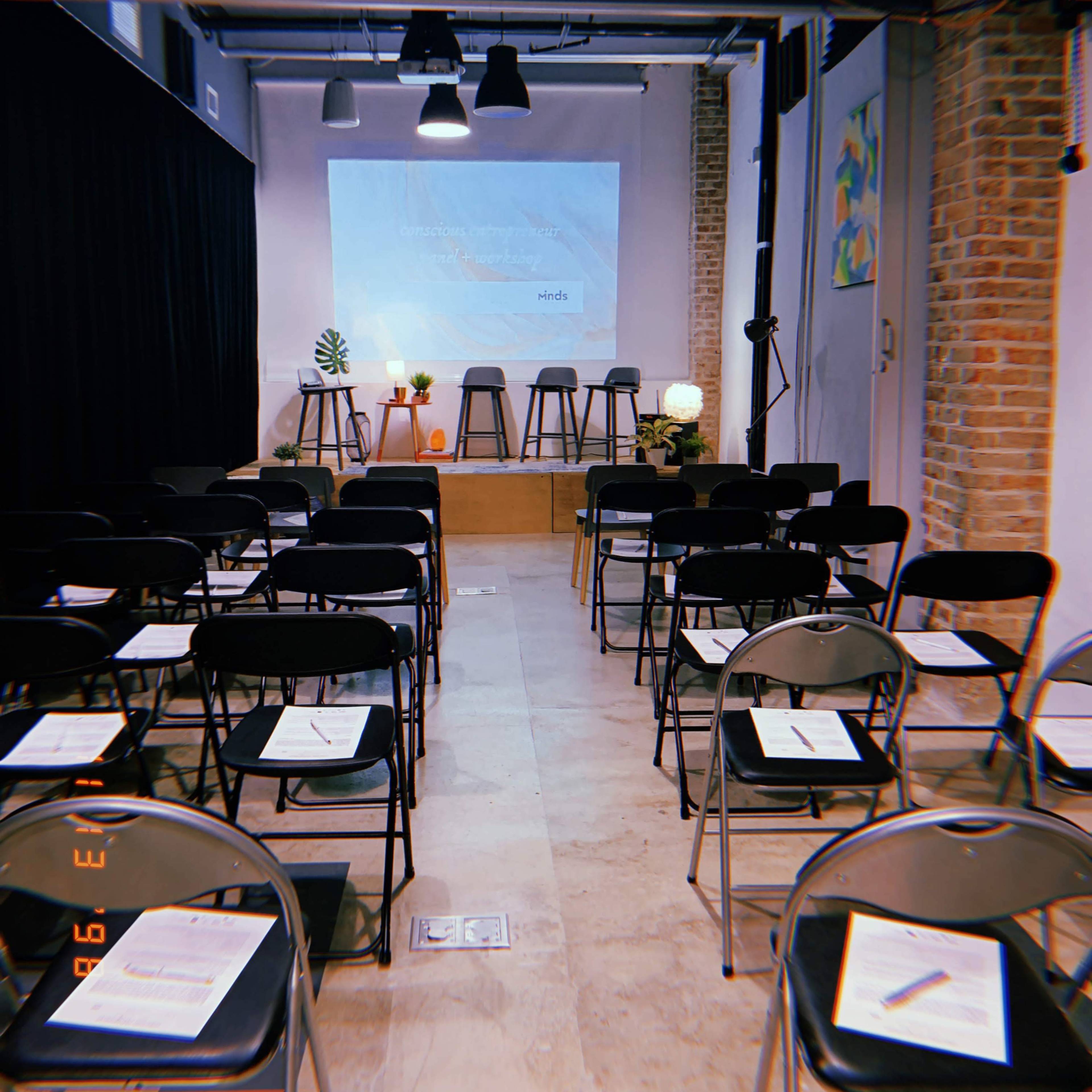An empty event space features rows of black chairs facing a small stage with a projector screen, surrounded by plants and decorative elements.