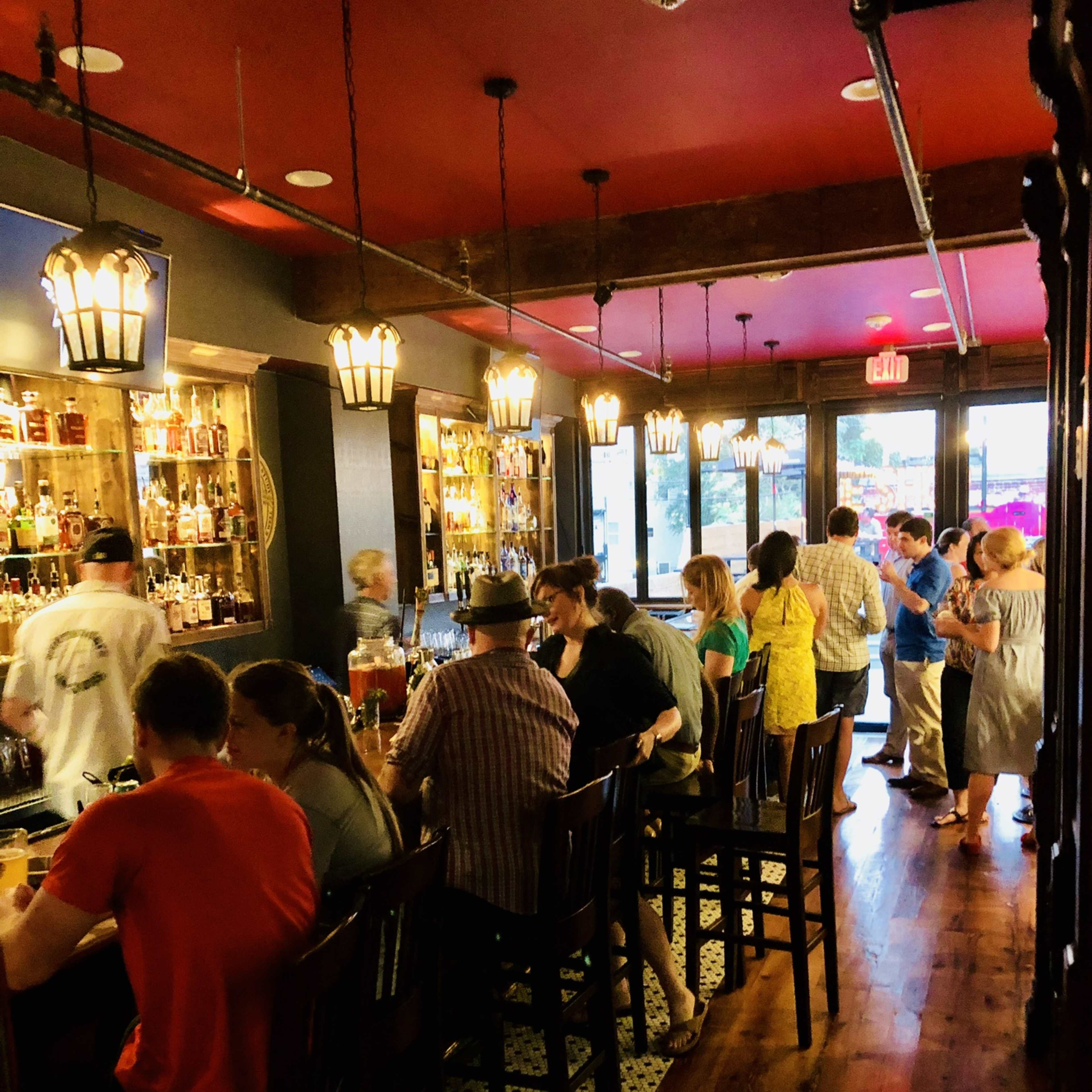 A busy bar interior with patrons seated at a long counter and others standing near shelves filled with bottles.