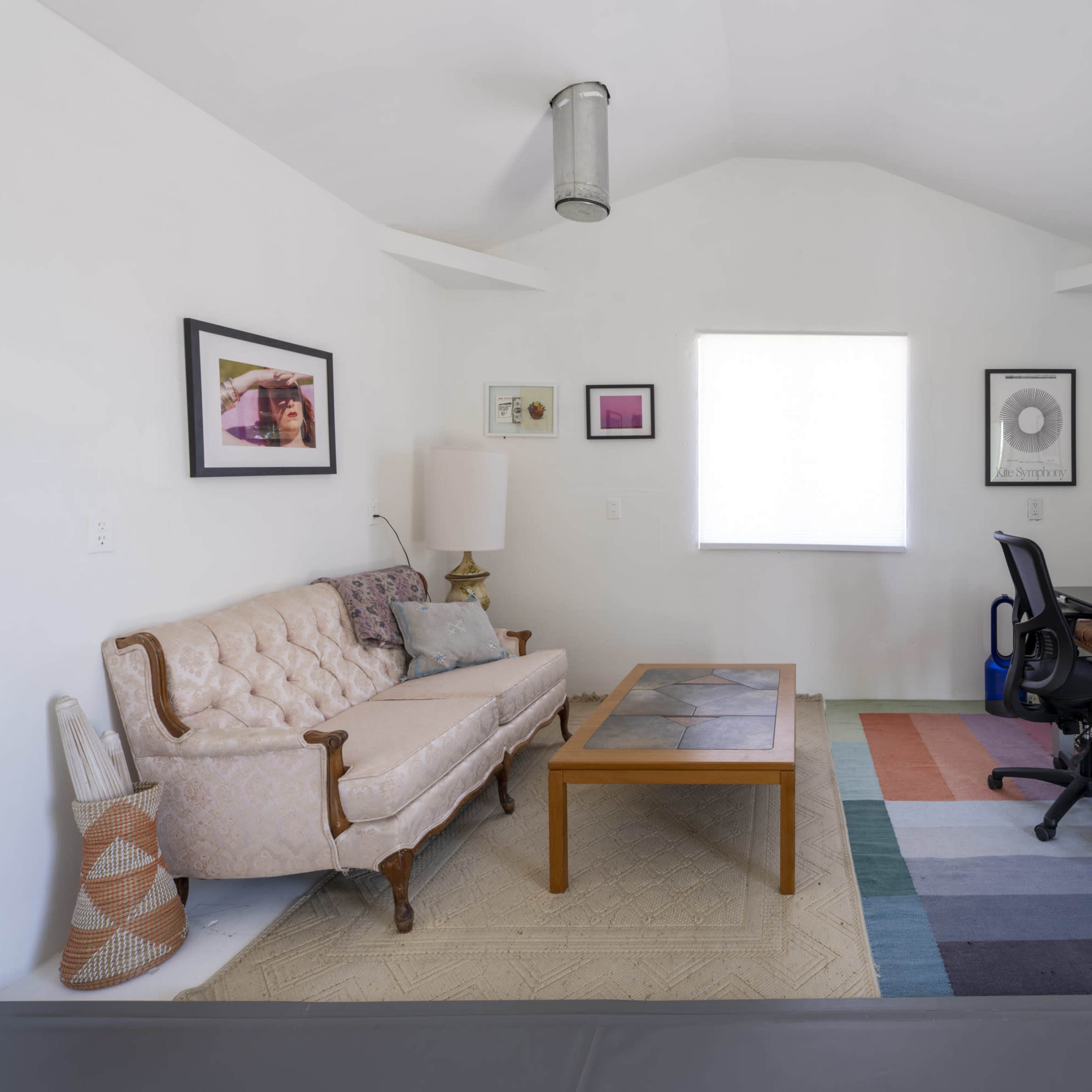 The room features a light-colored interior with a sofa, coffee table, and a desk with a chair, complemented by various framed artworks on the walls.