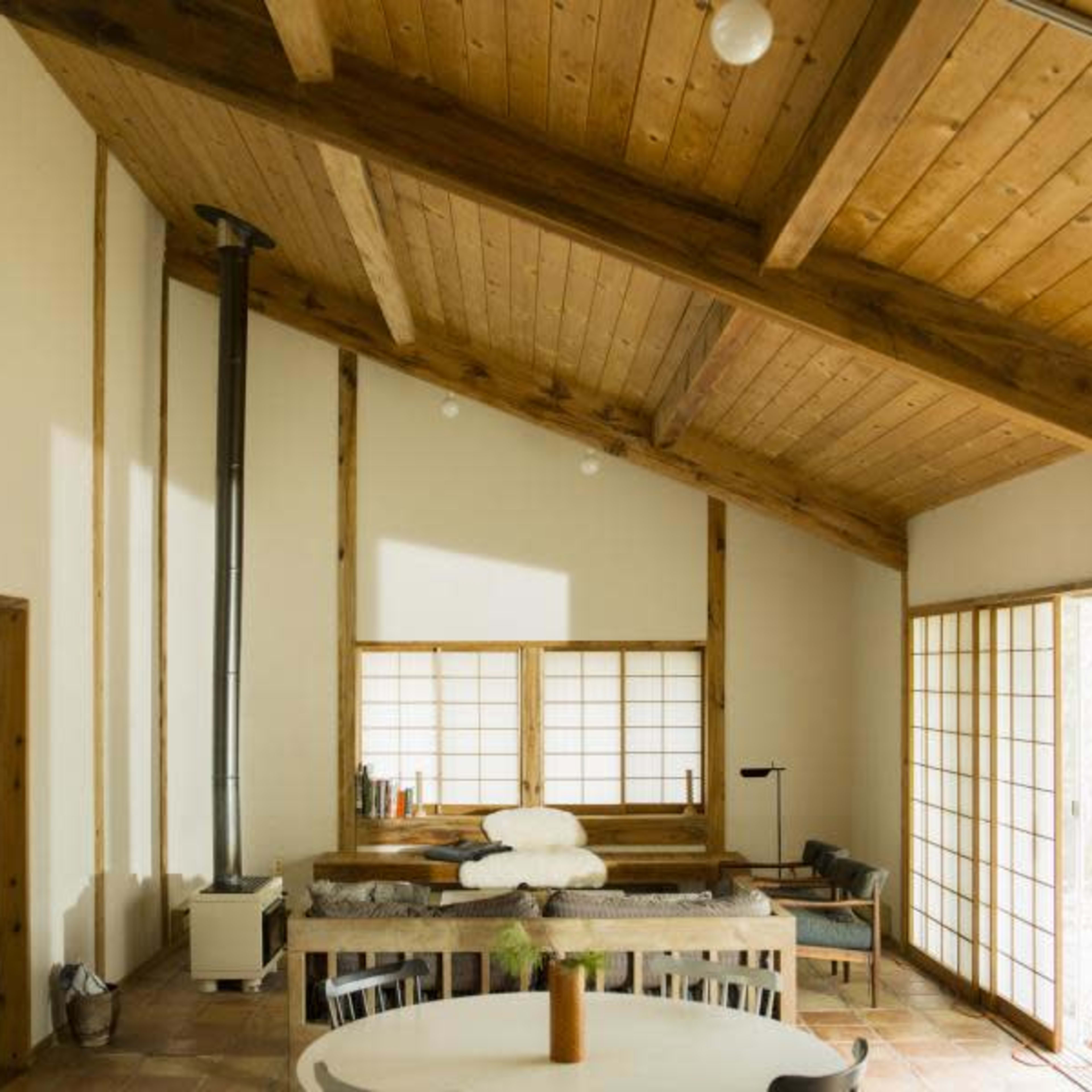 A modern interior of a house with wooden beams, a central dining table, and large windows that open to a natural setting.