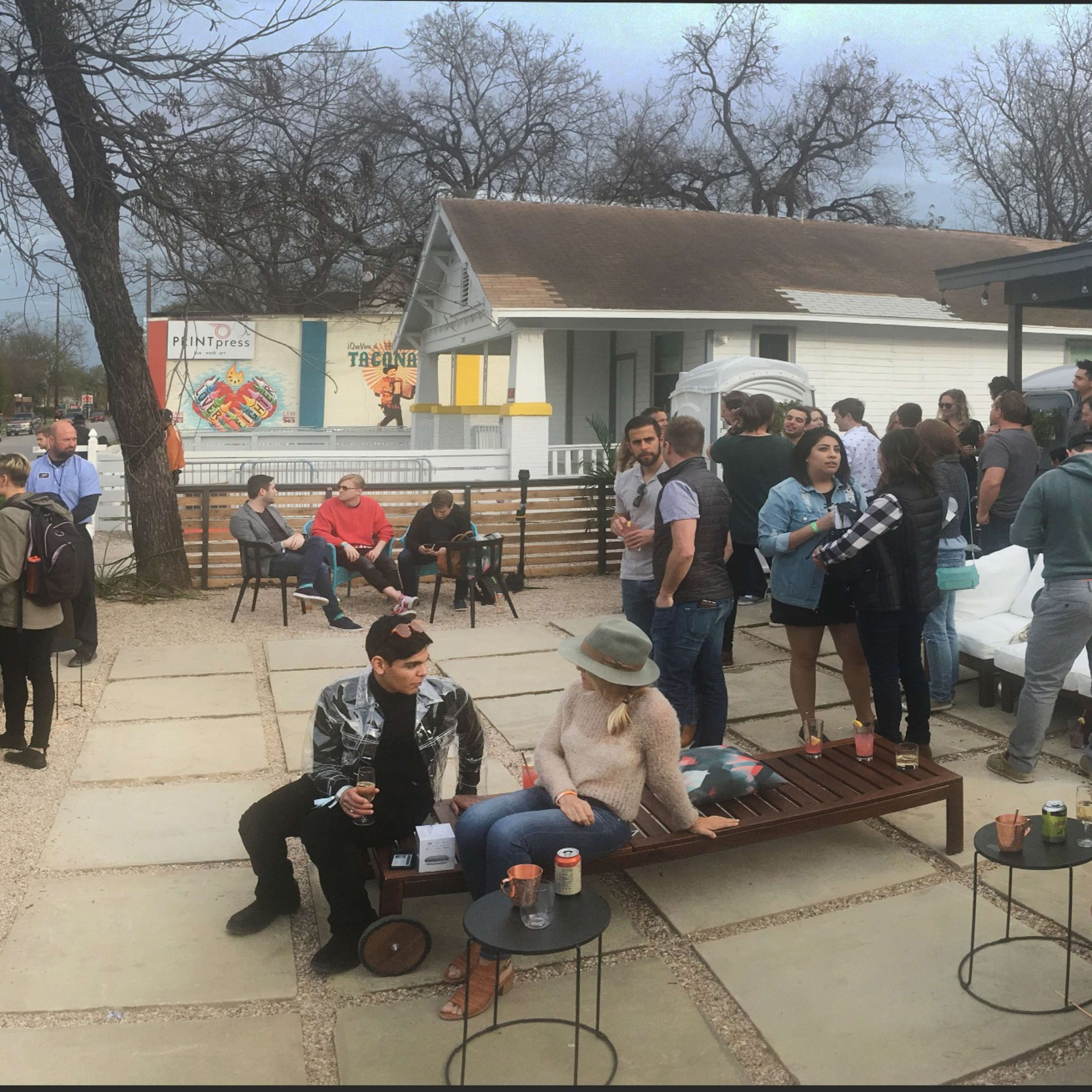 A diverse group of people socializes outside a restaurant with a spacious patio, surrounded by trees and a cloudy sky.