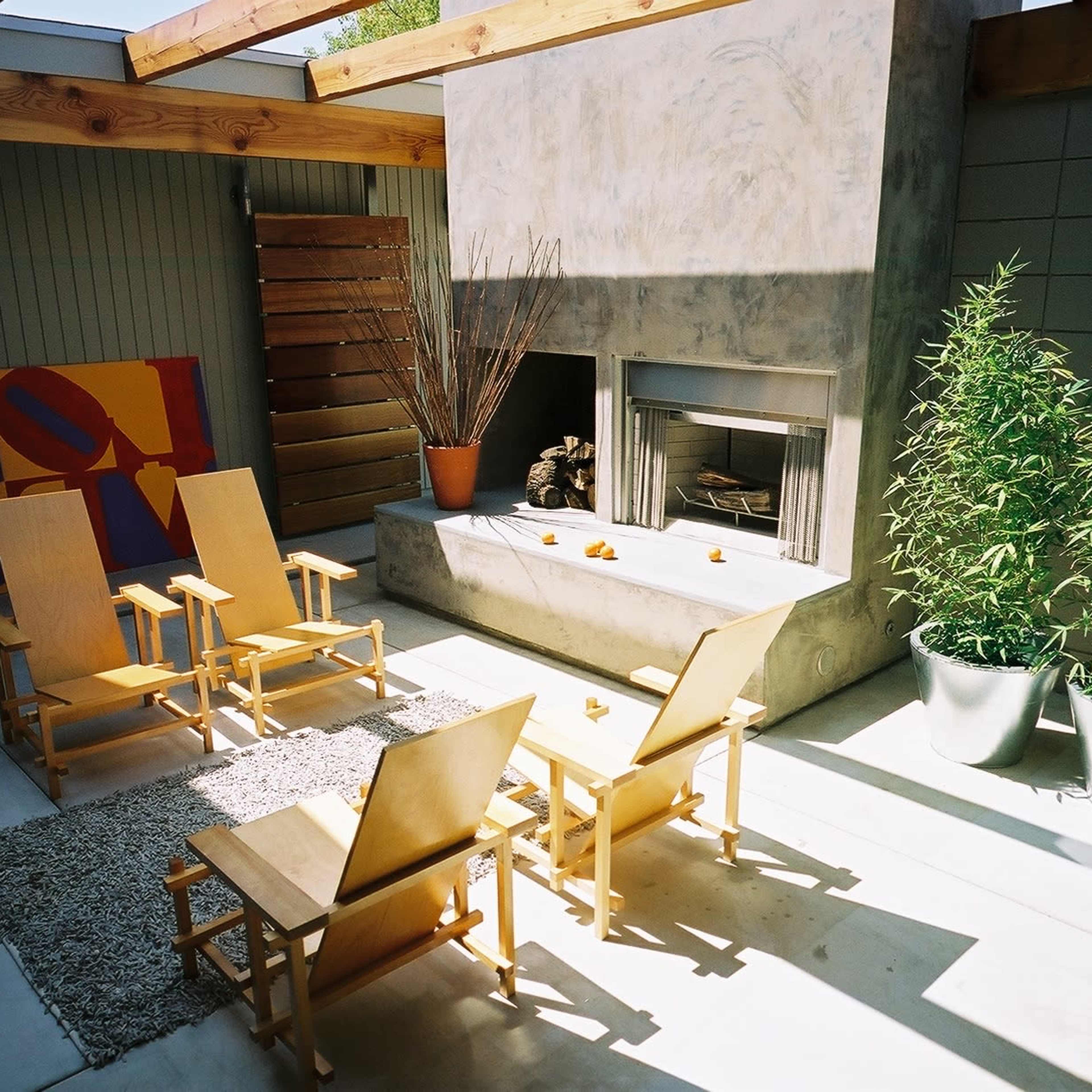 A modern outdoor seating area with four wooden chairs arranged around a concrete fireplace, surrounded by potted plants and a textured rug on the floor.