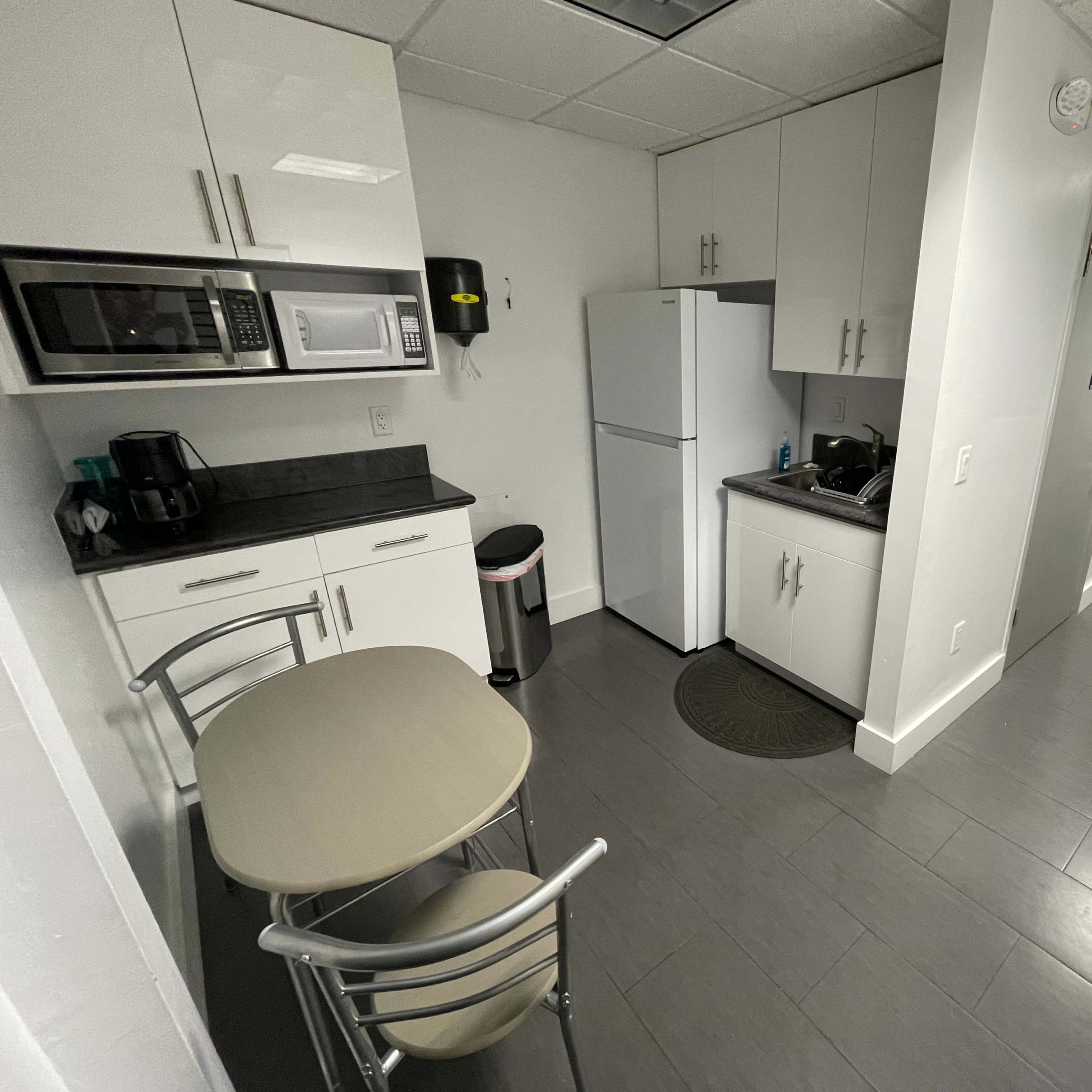A small kitchen area with a table and two chairs, a refrigerator, a microwave, and a sink, set in a modern, minimalist design.