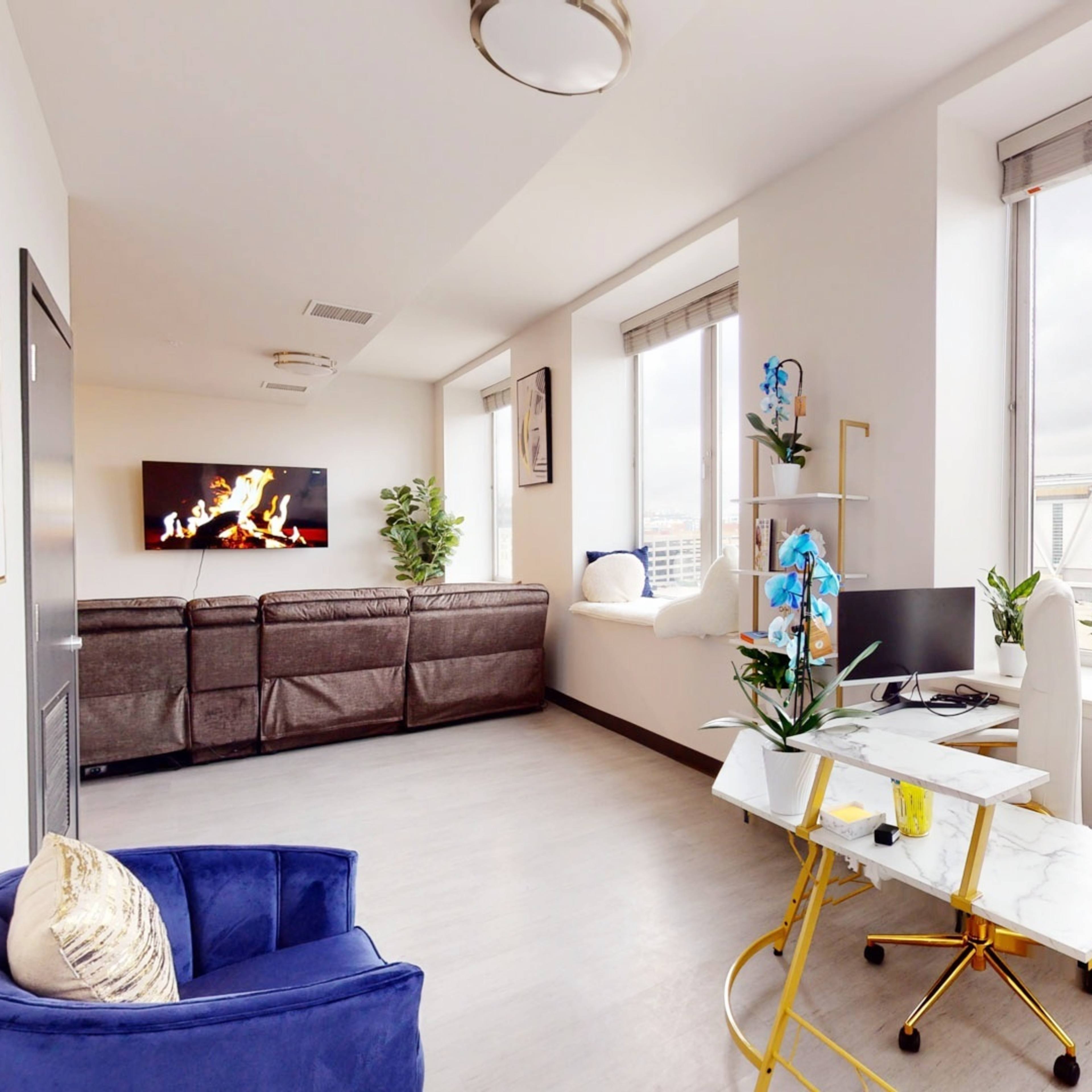 A modern living room features a blue chair, a small desk with plants, and large windows overlooking the city.