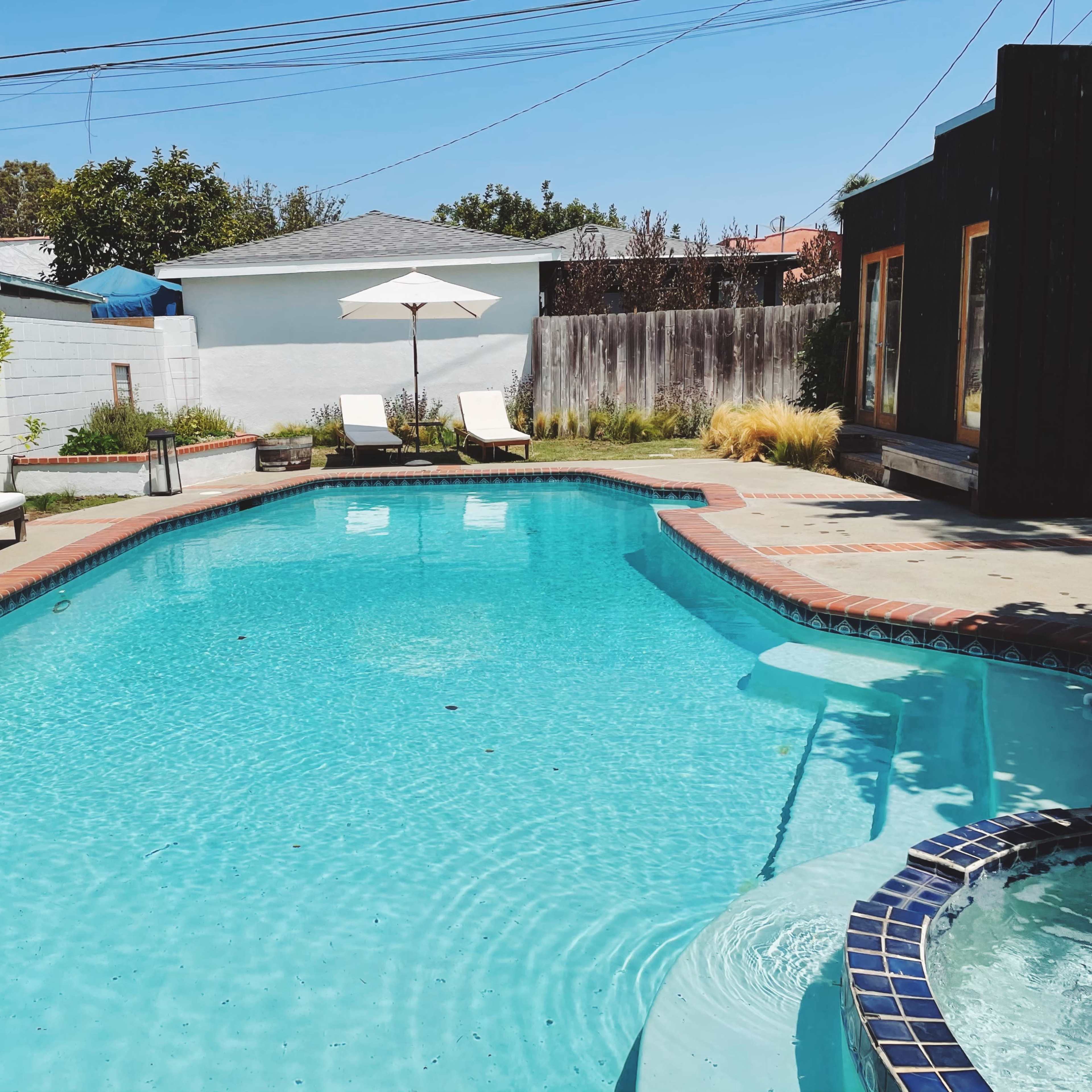 A clear blue swimming pool is surrounded by lounge chairs and a patio, with a shaded umbrella in the background and a wooden structure nearby.