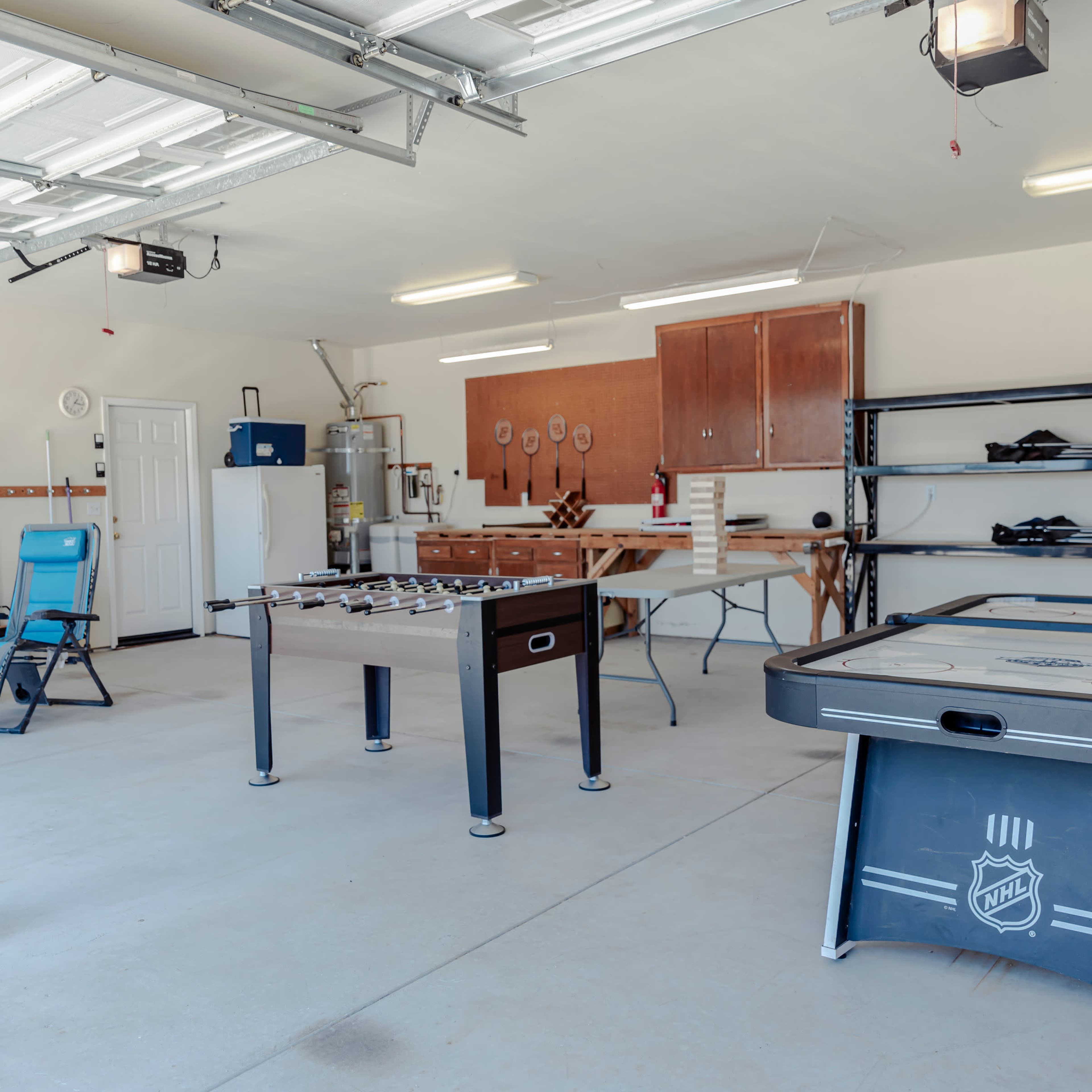 A spacious garage equipped with a foosball table, an air hockey table, a worktable, shelves, and two folding chairs.