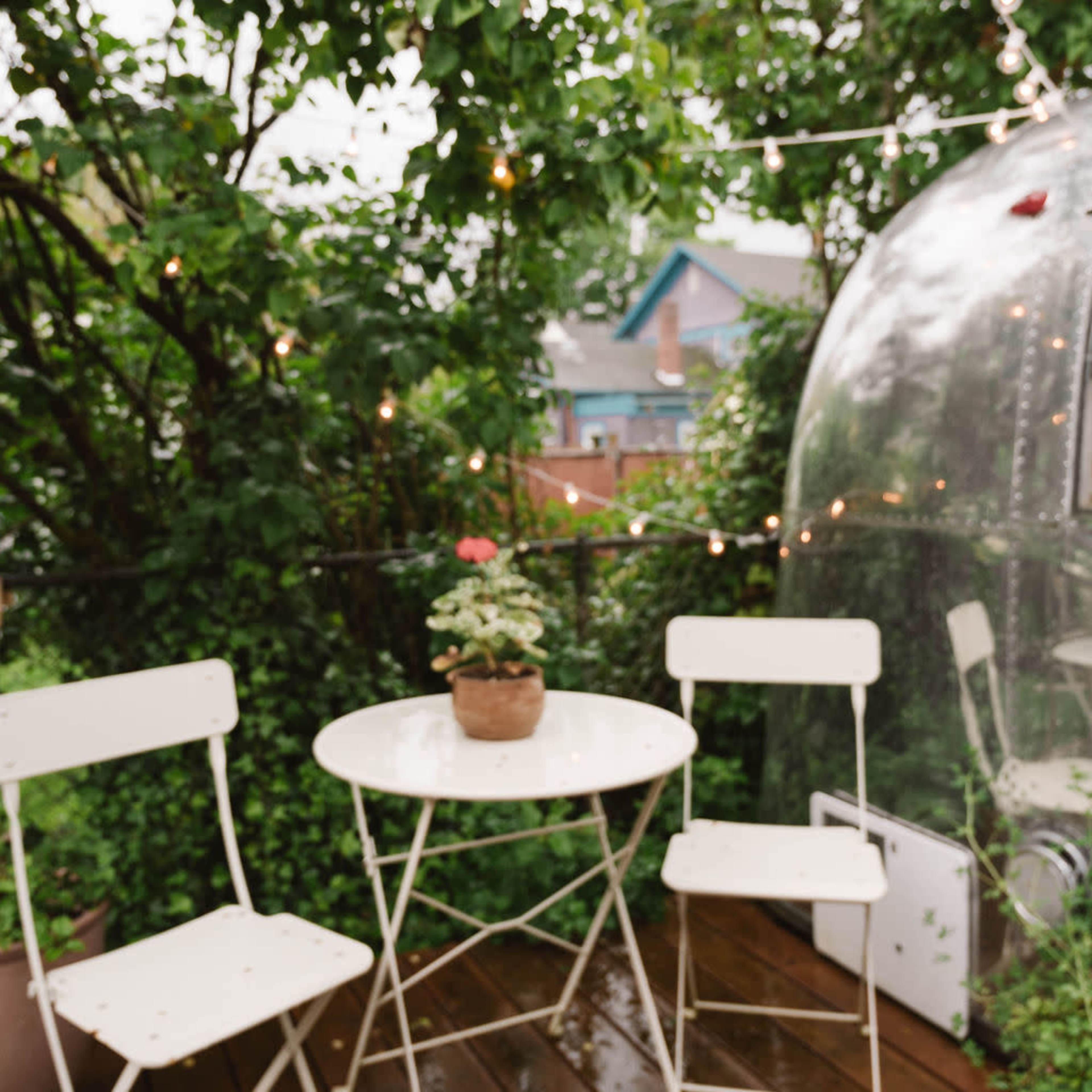 A small outdoor seating area features a round table with a potted plant and two white folding chairs, surrounded by greenery and string lights.