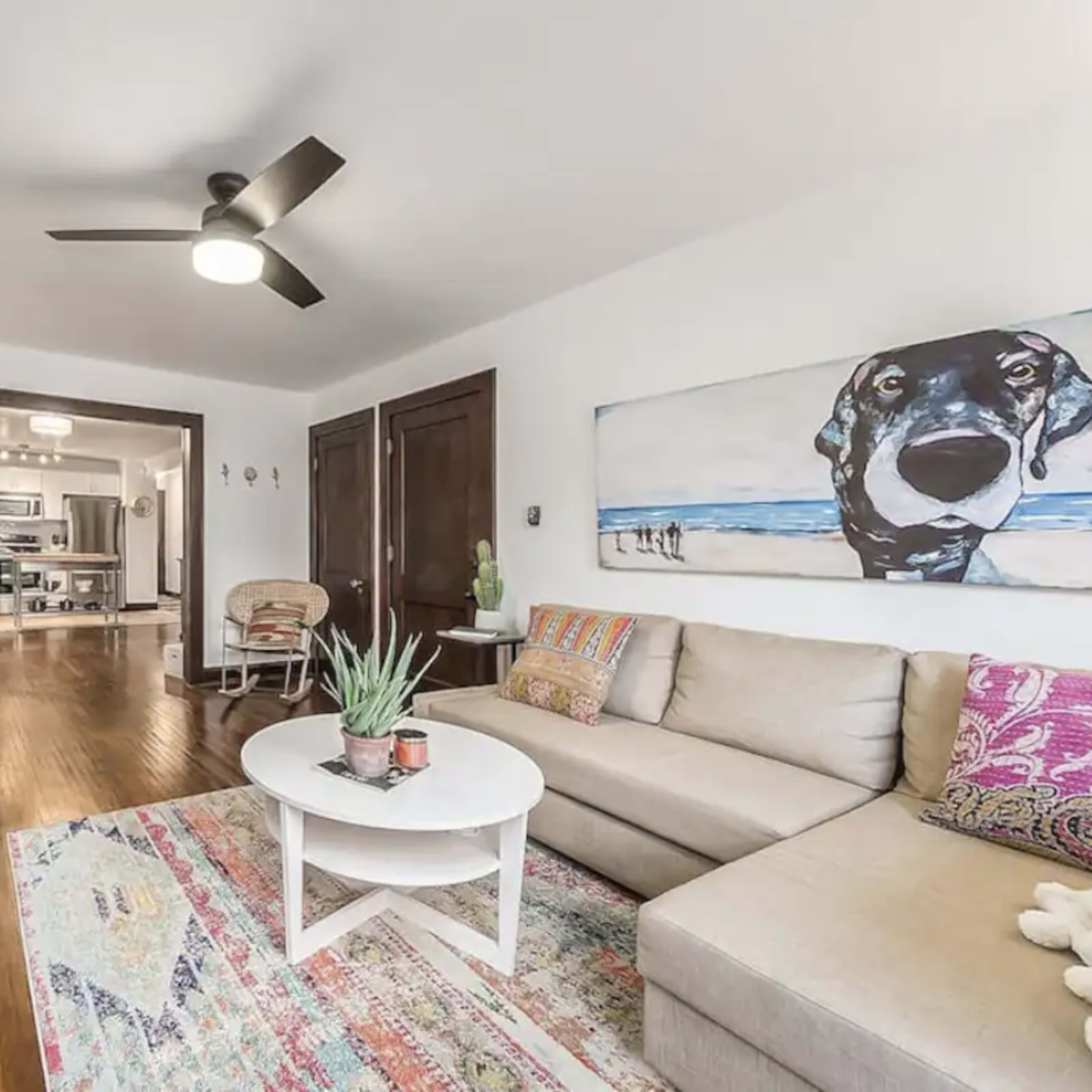 A living room with a beige sectional sofa, a round coffee table, a decorative rug, a wall-mounted television, and a large painting of a dog.