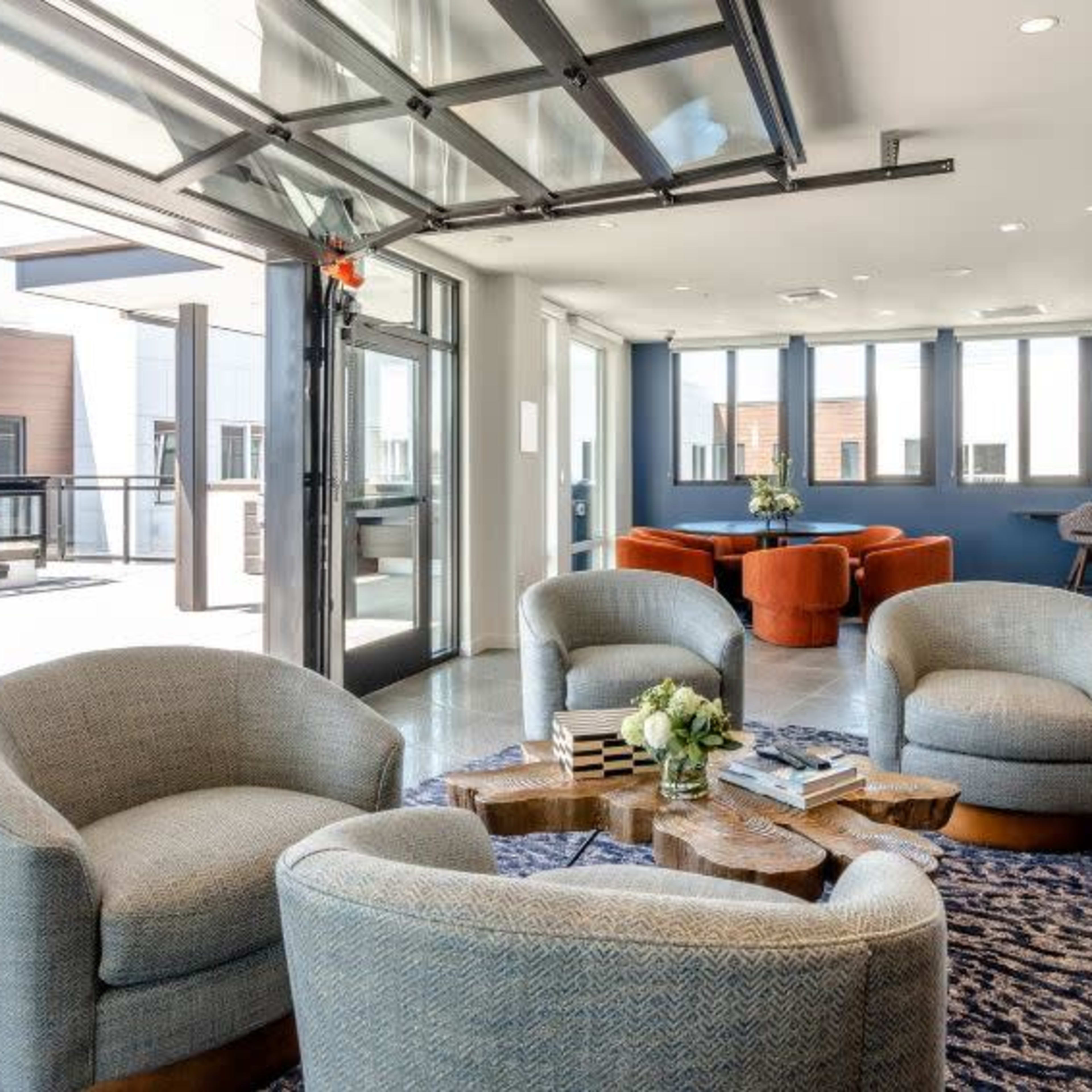 A modern lounge area with four curved chairs around a wooden coffee table, large windows, and a glass garage door that opens to an outdoor space.