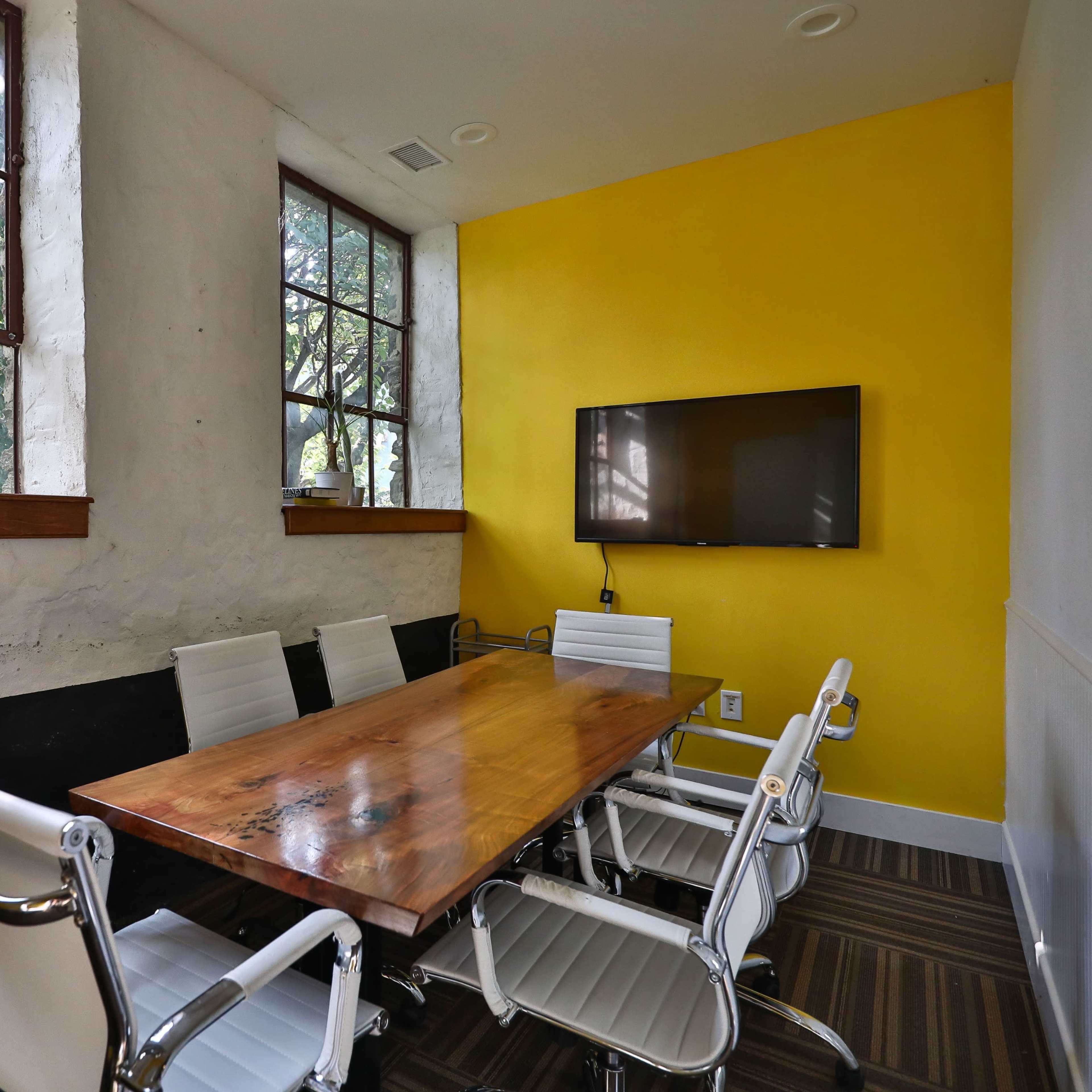 A small conference room features a wooden table surrounded by white chairs, set against a yellow wall and large windows.