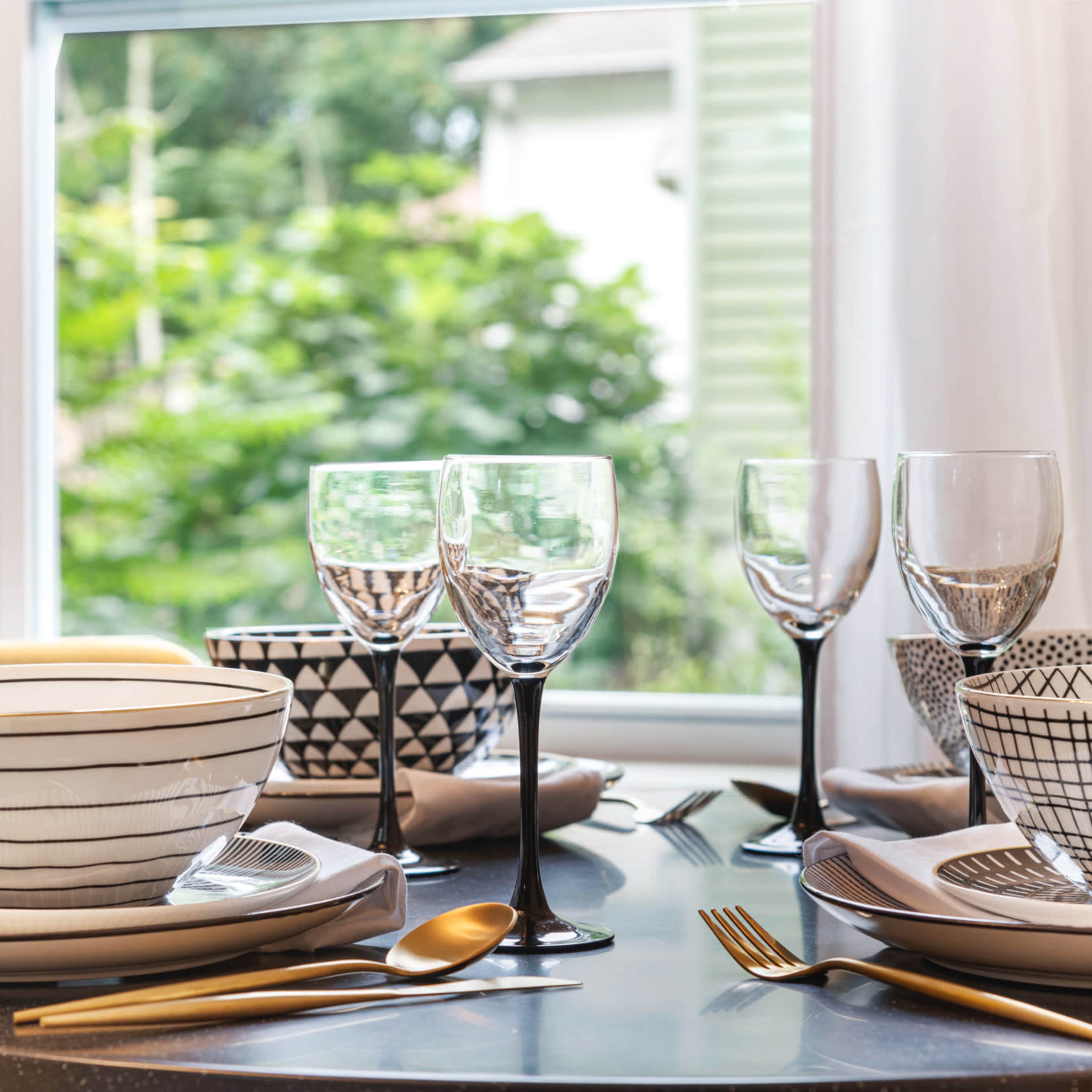 A dining table is set with patterned bowls, wine glasses, and gold utensils, with a window displaying greenery in the background.
