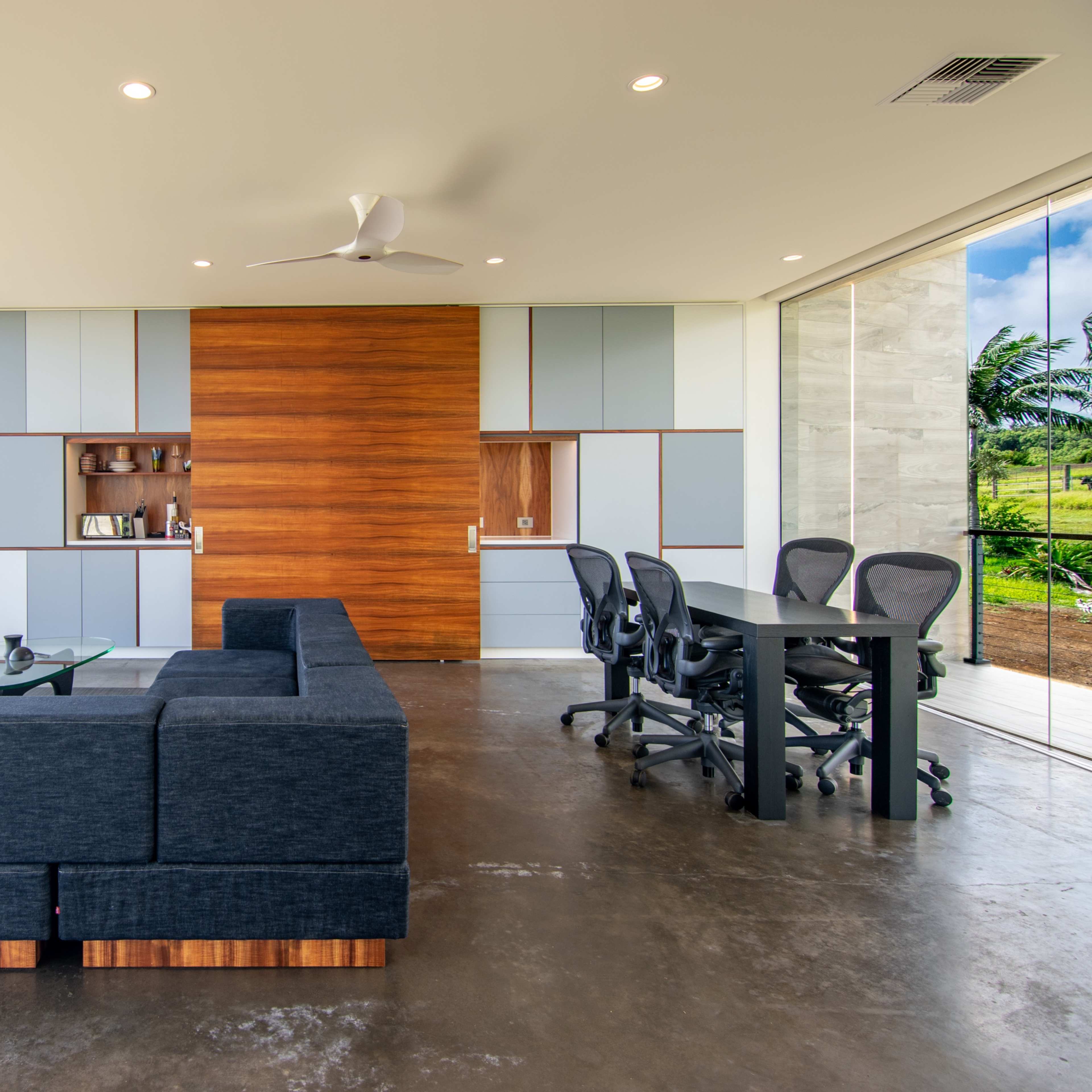 A modern open-concept living area featuring a dark sectional sofa, a wooden accent wall, a glass table, and a dining set, with large windows revealing a view of greenery and a cloudy sky.