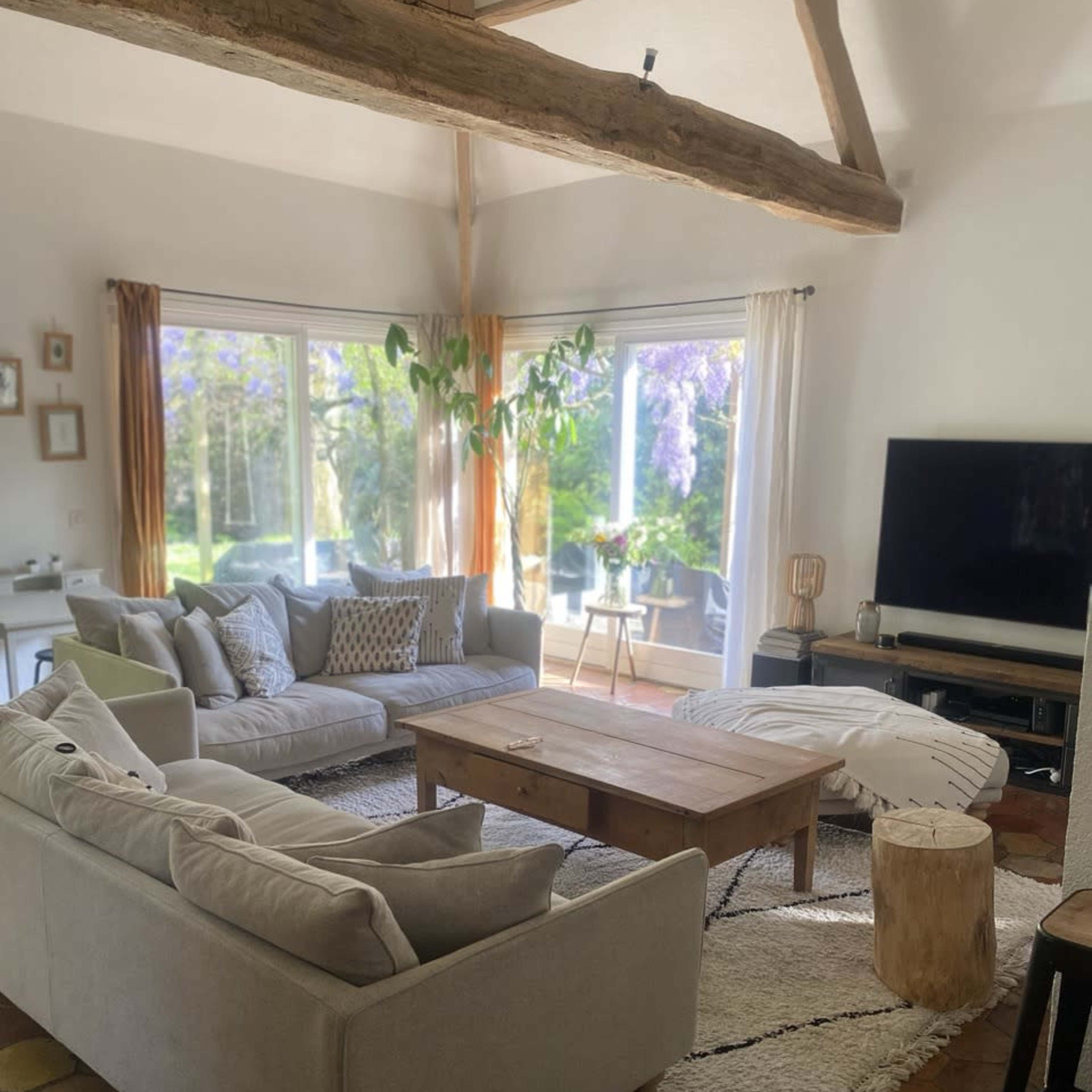 A cozy living room with two light-colored sofas arranged around a wooden coffee table, featuring a large window that opens to a green outdoor space.