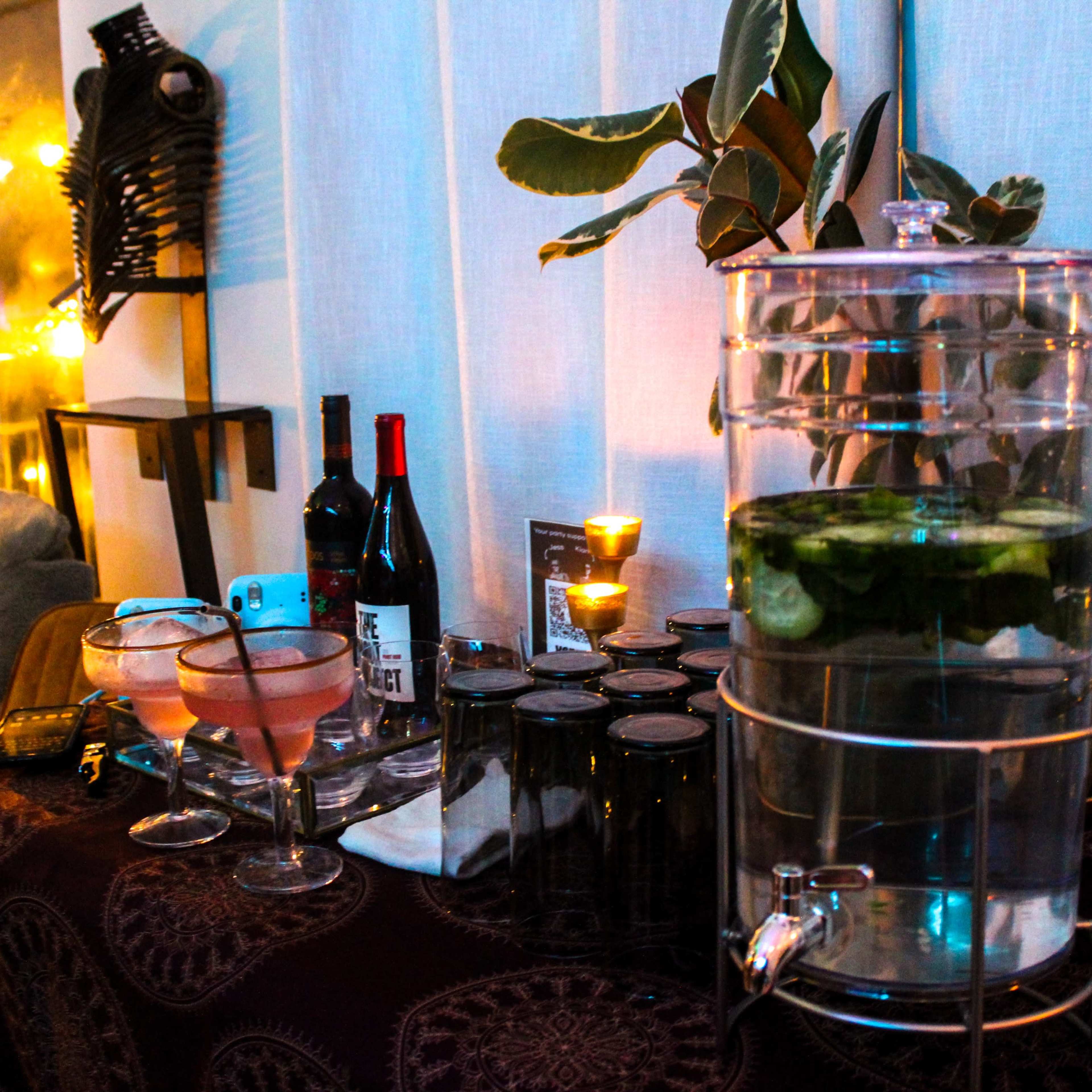 A table is set with a drink dispenser filled with water and cucumbers, alongside bottles of wine, glasses, and candles in a dimly lit room.