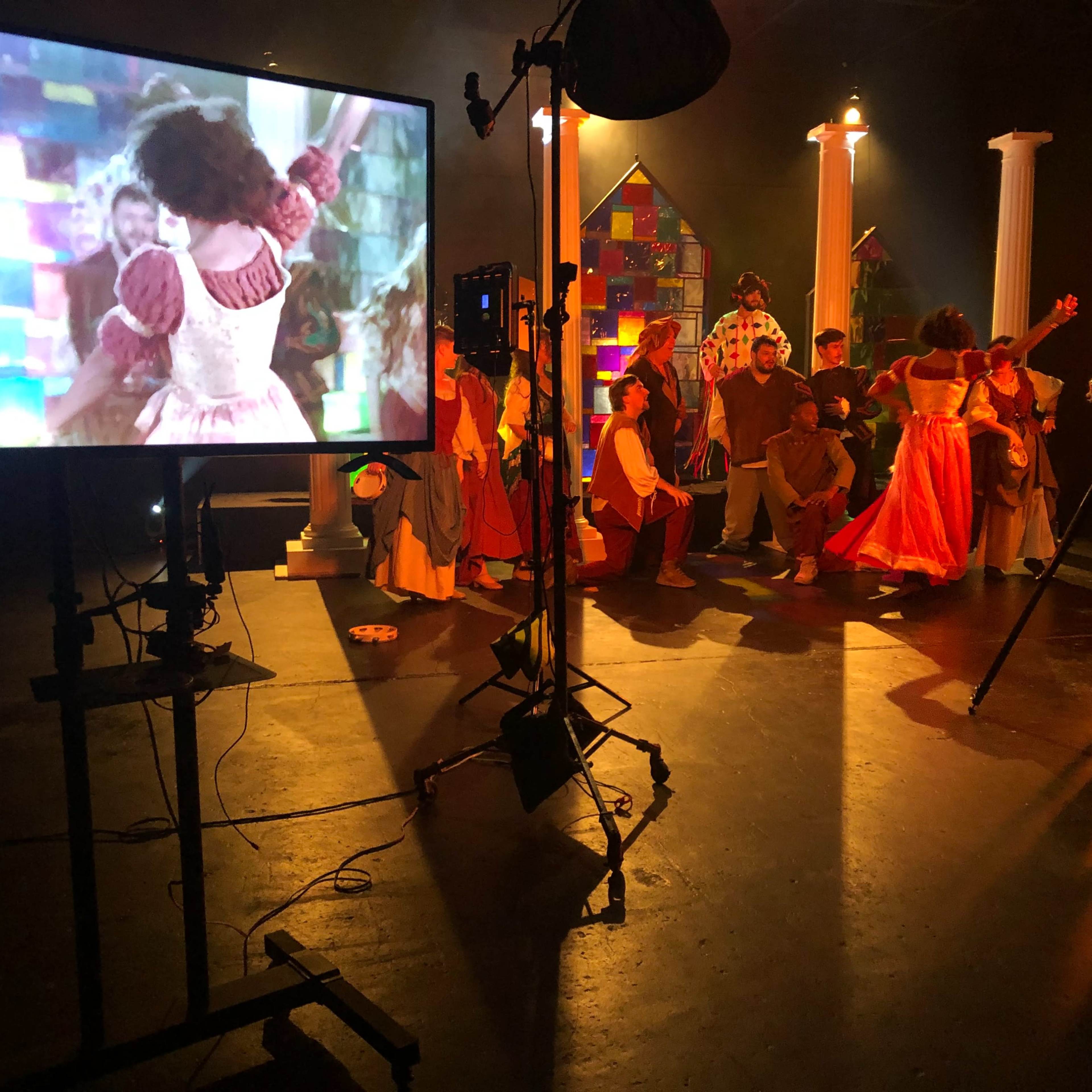 A film set features a large monitor displaying a scene, while actors in colorful costumes gather and perform in front of it.