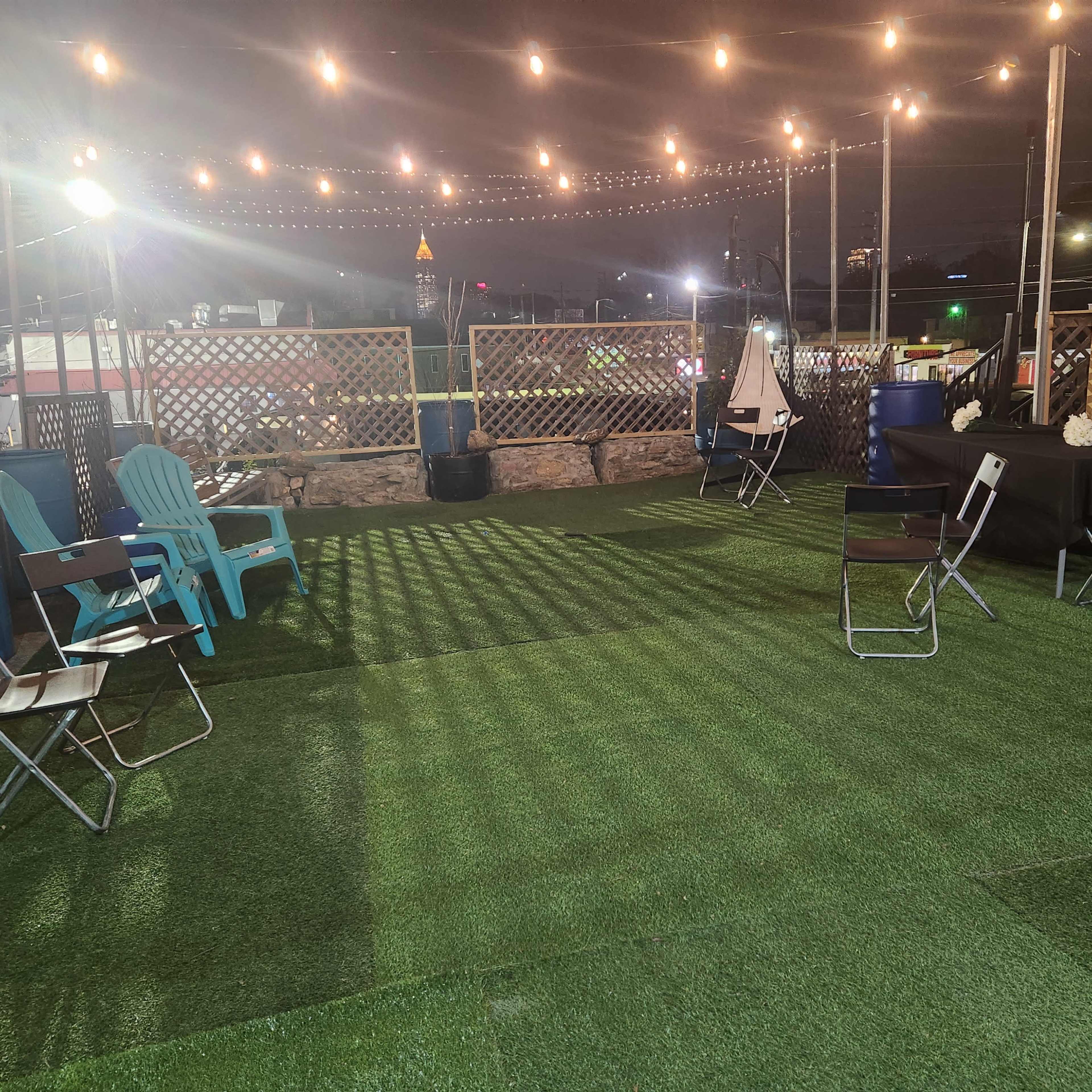 An outdoor gathering space features green artificial turf, string lights overhead, and a mix of seating options including chairs and tables.