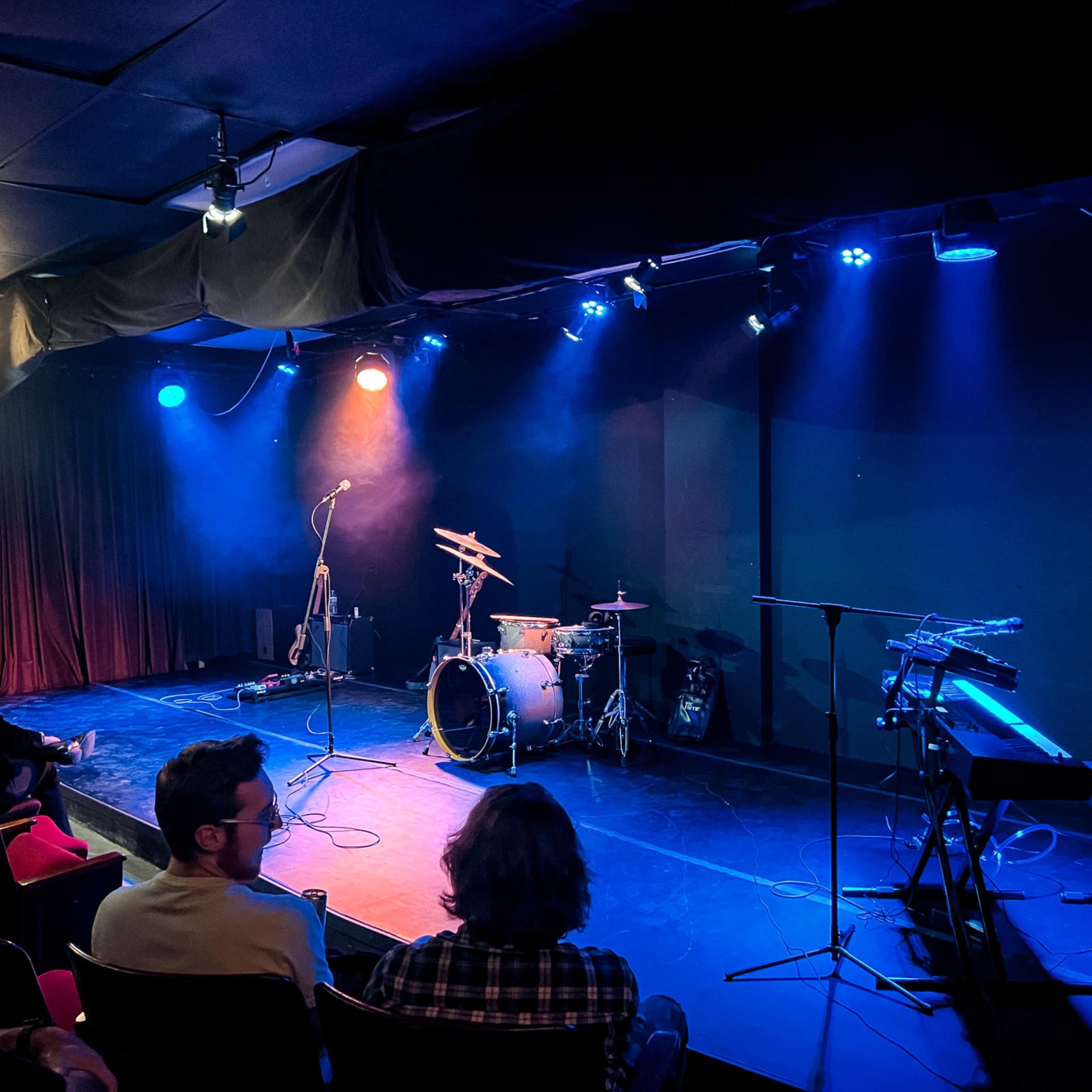 The stage is set for a performance, featuring a drum kit, a keyboard, and various instruments under blue and orange lights, with a small audience seated nearby.