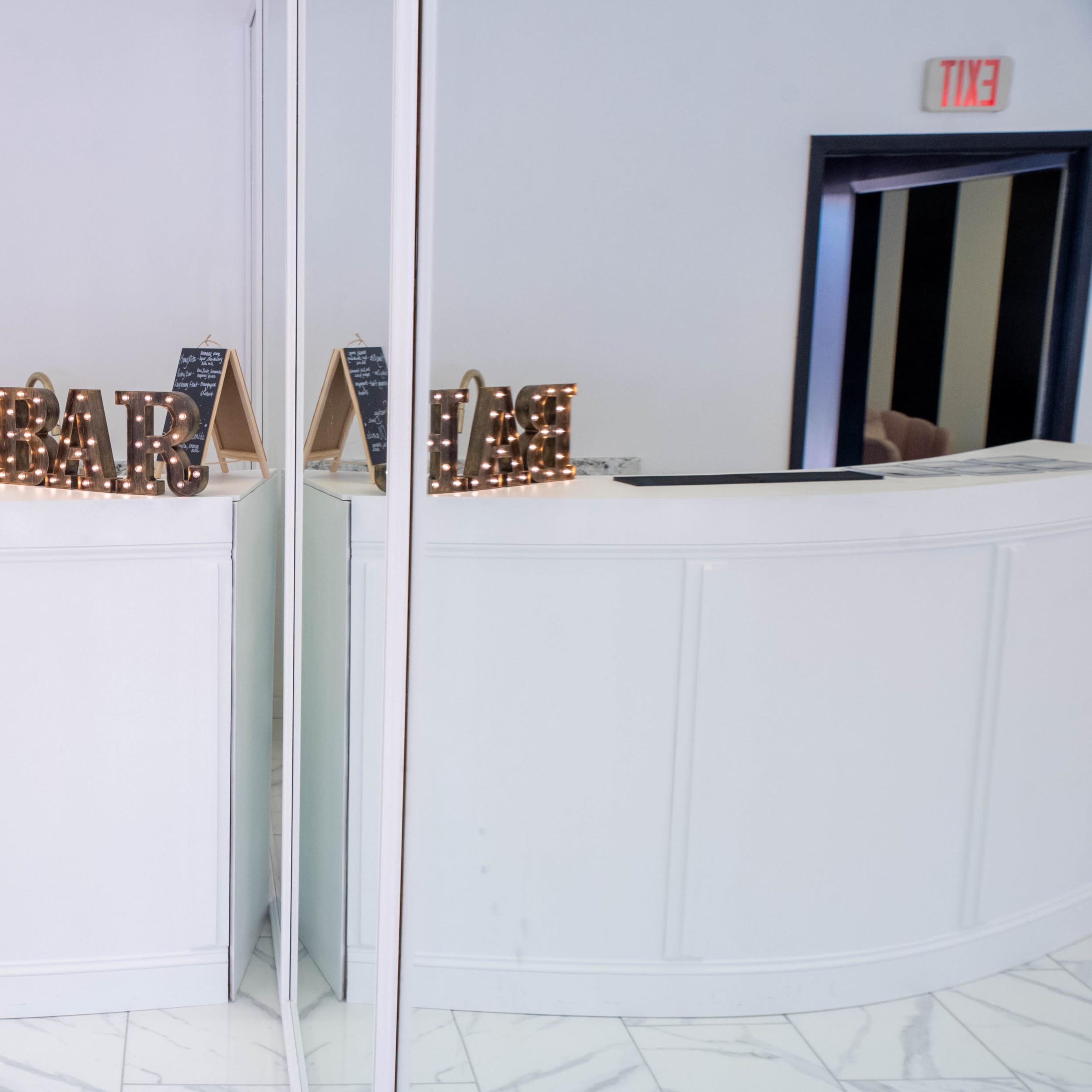 A curved white reception desk with illuminated letters spelling "BAR" is mirrored in a spacious area with exit signs above.